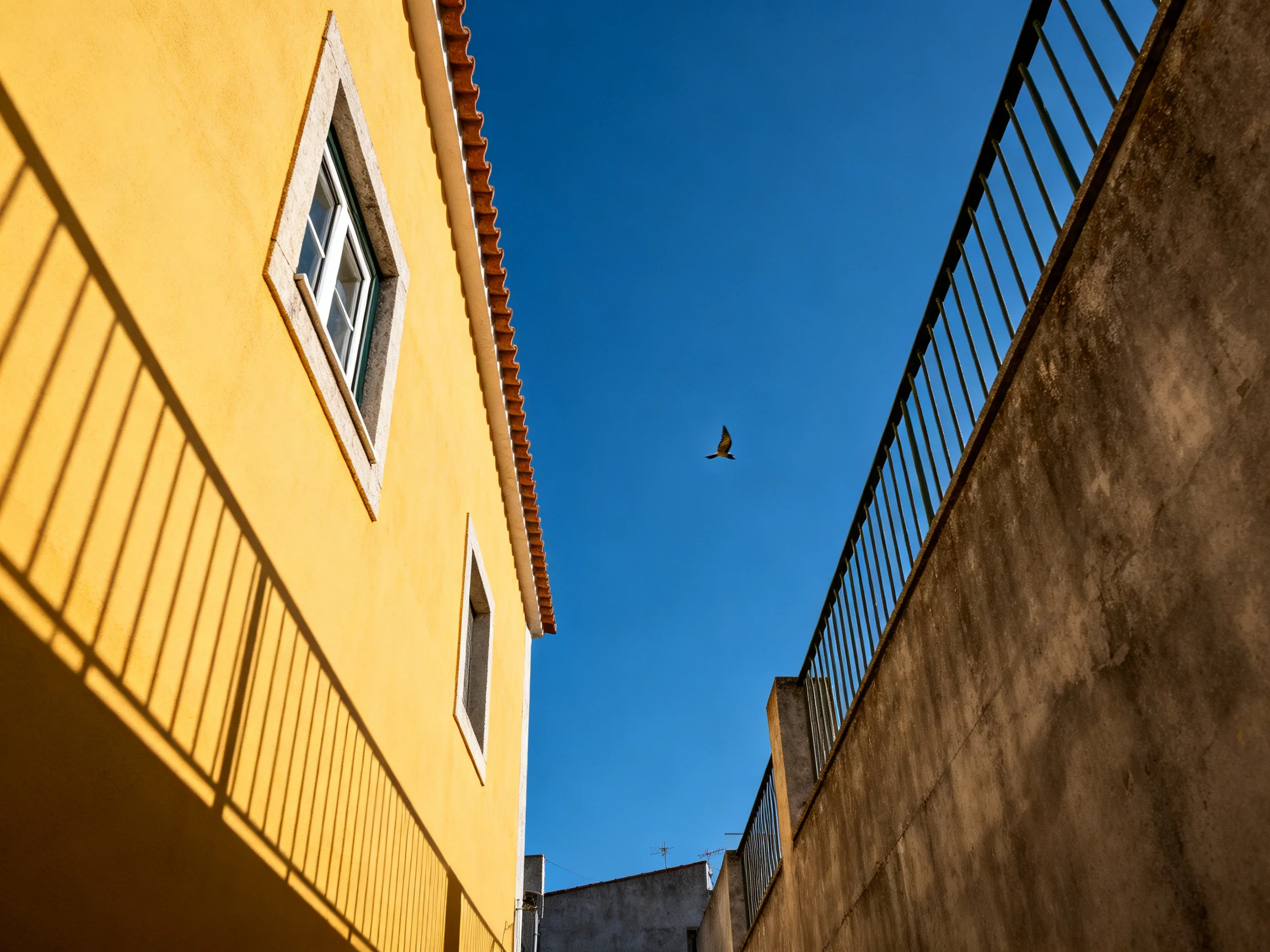 Narrow alley between yellow building and stone wall with blue sky and flying bird