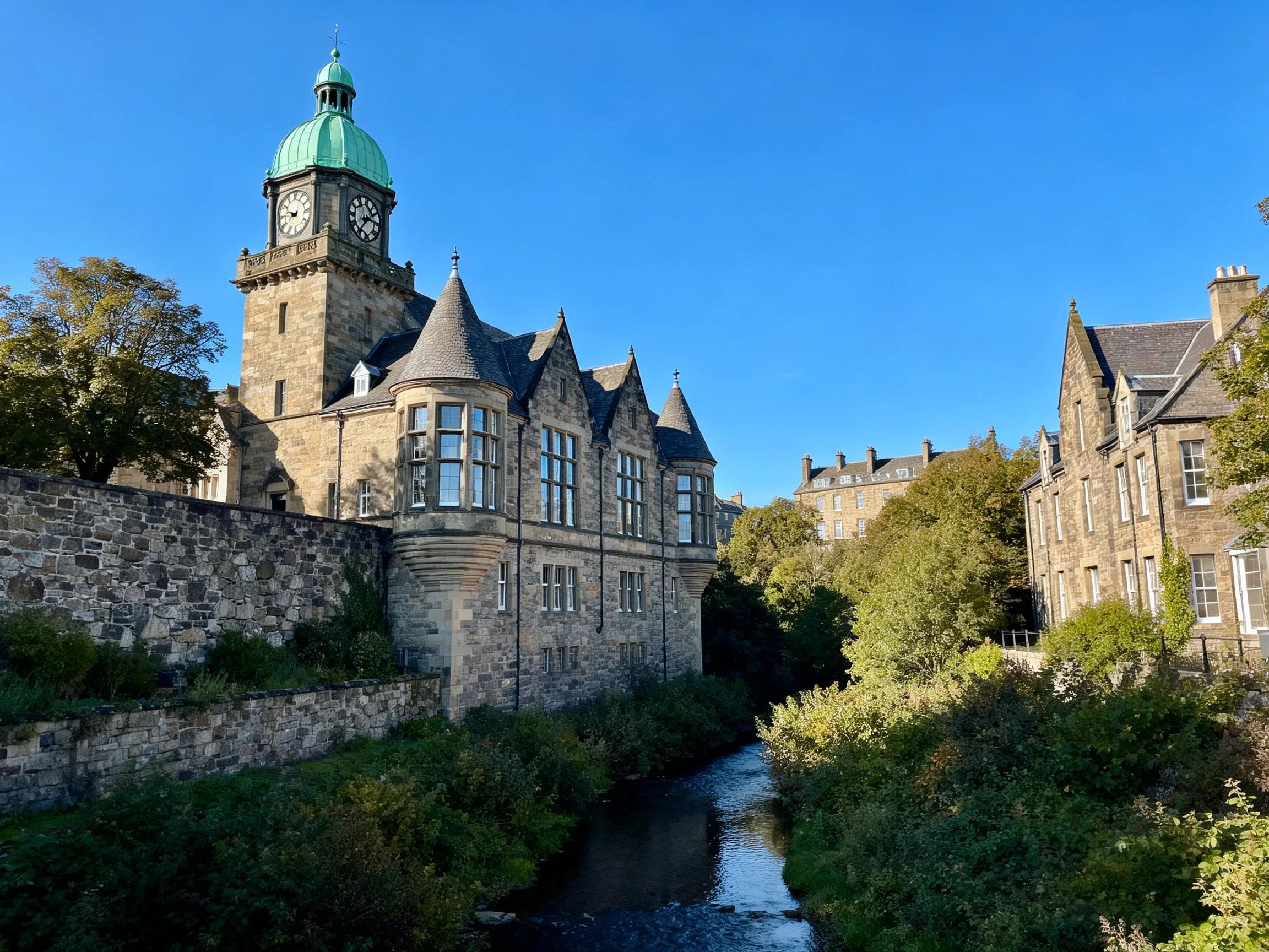 A historic stone building with a green-domed clock tower near a stream