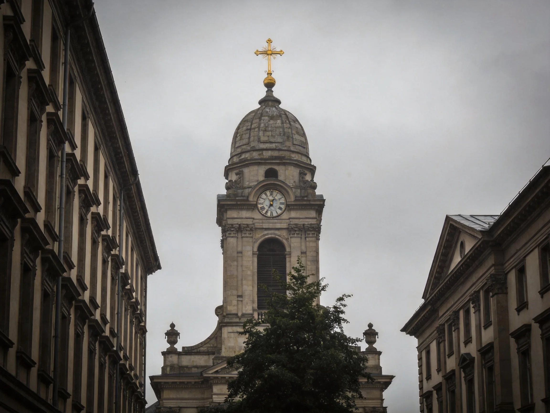 A historic stone church tower with golden cross and clock, flanked by old buildings