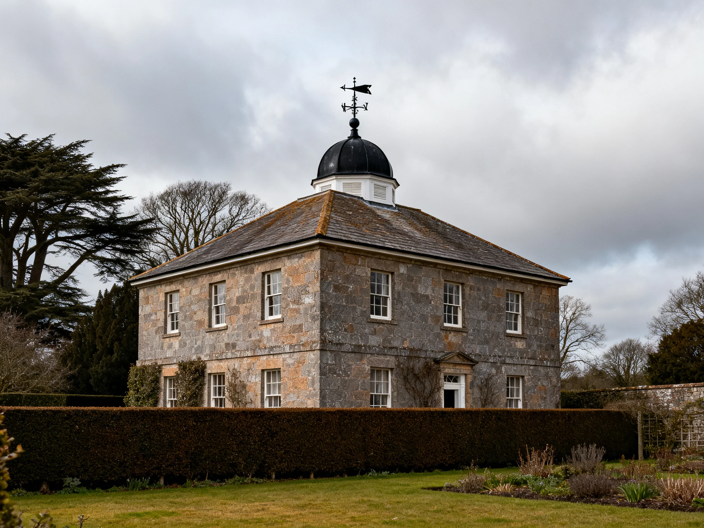 A historic stone house with a domed roof and weather vane near manicured gardens