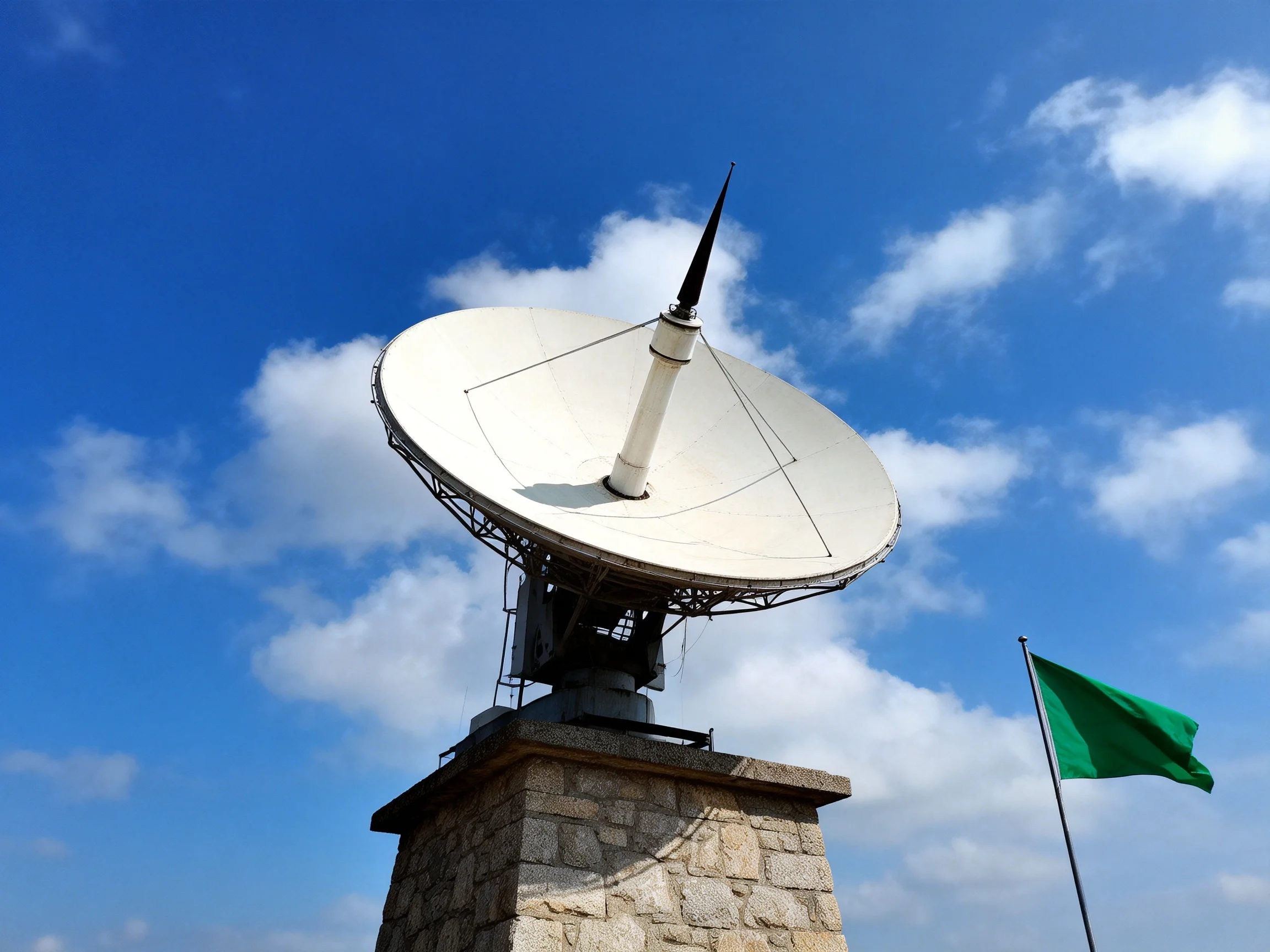 A large white satellite dish on a stone pedestal near a green flag under blue sky