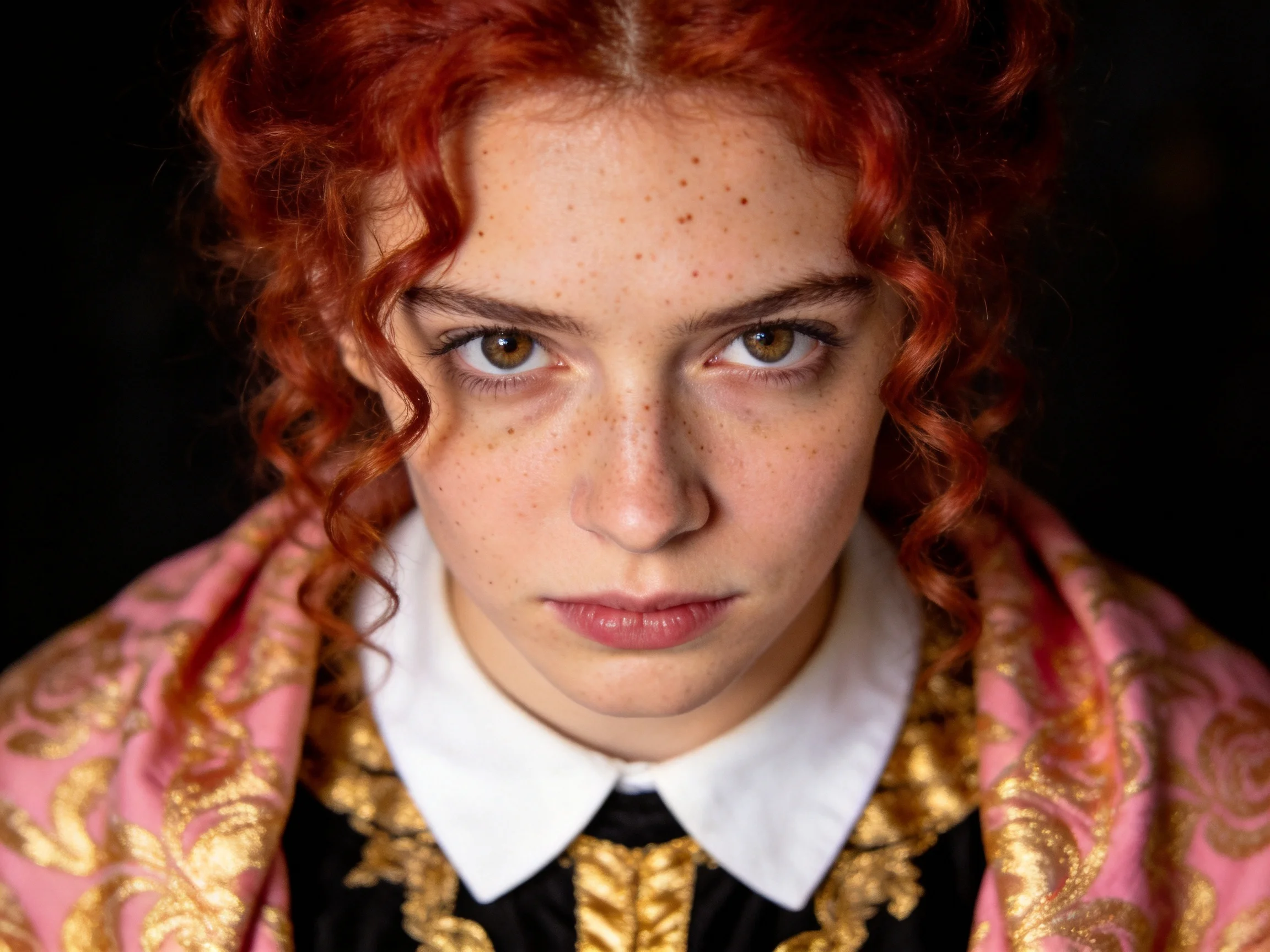 A person with vibrant red curly hair wearing ornate pink and gold garments