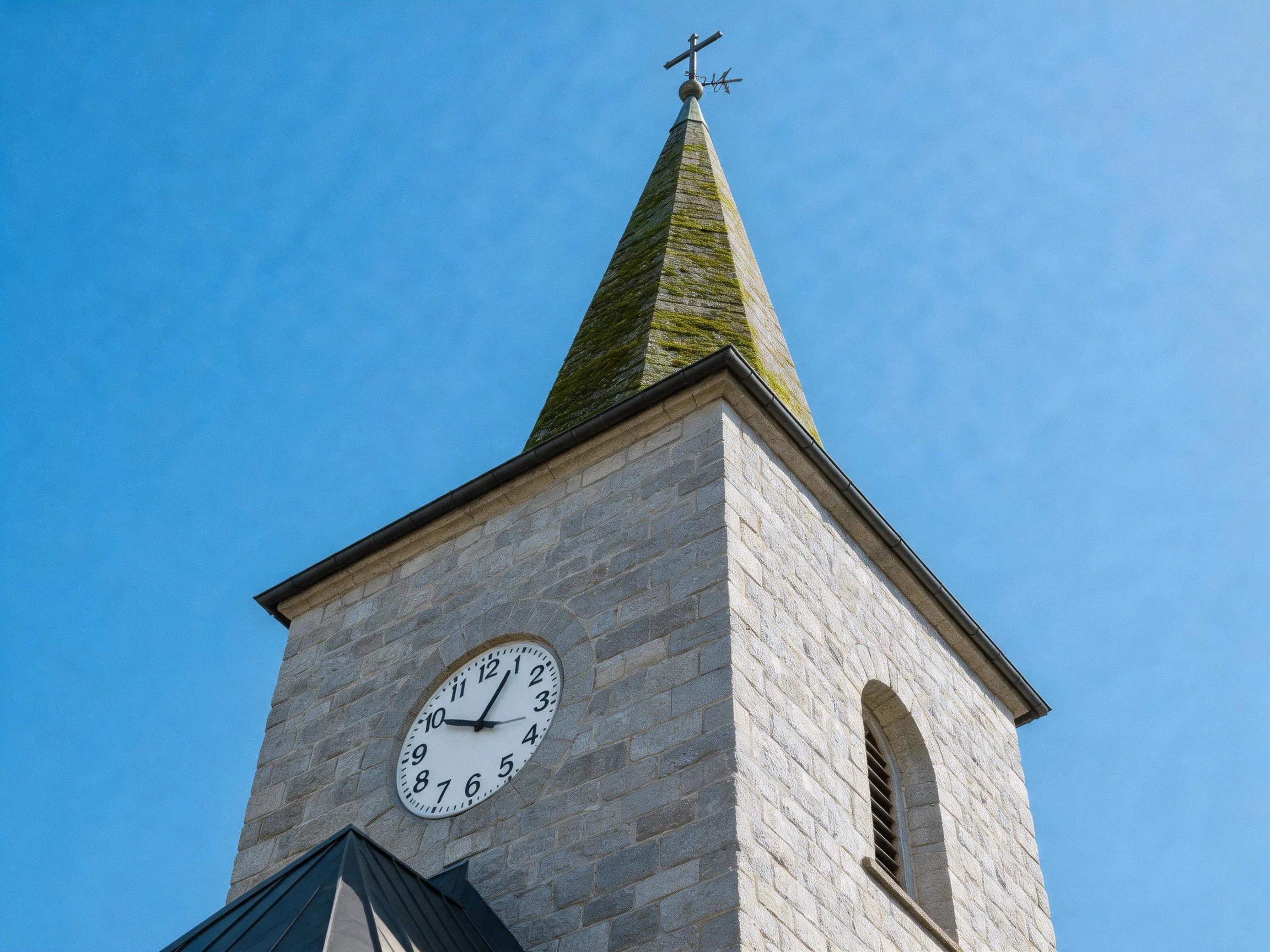 A stone church tower with a clock and mossy spire under clear blue sky