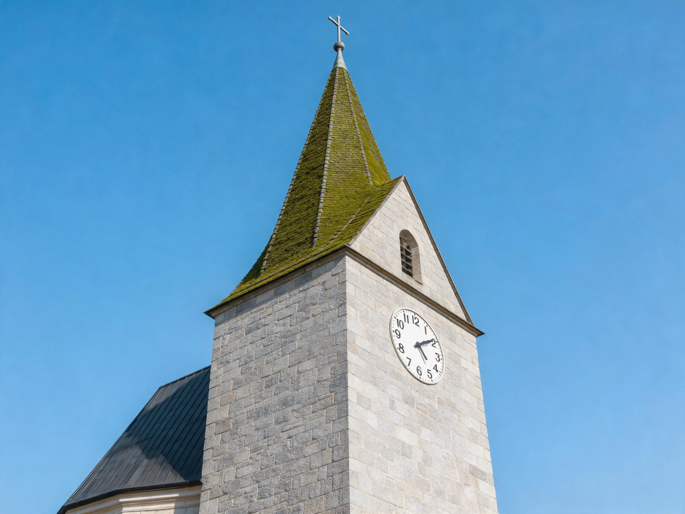A stone church tower with a moss-covered spire and clock, set against a clear blue sky.