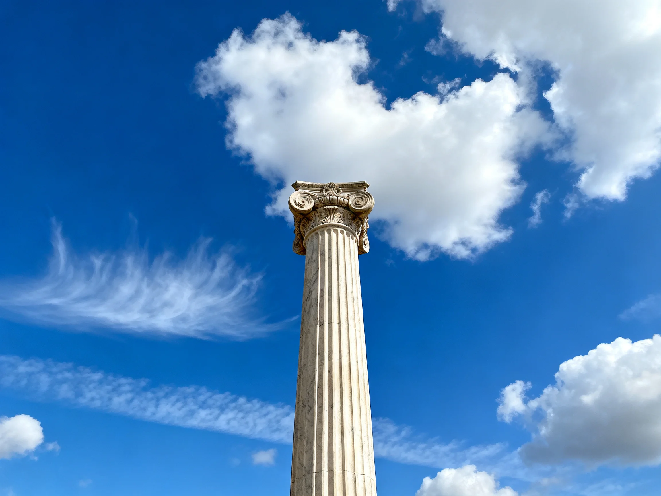 A tall ancient column with ornate capital beneath a bright blue sky filled with clouds