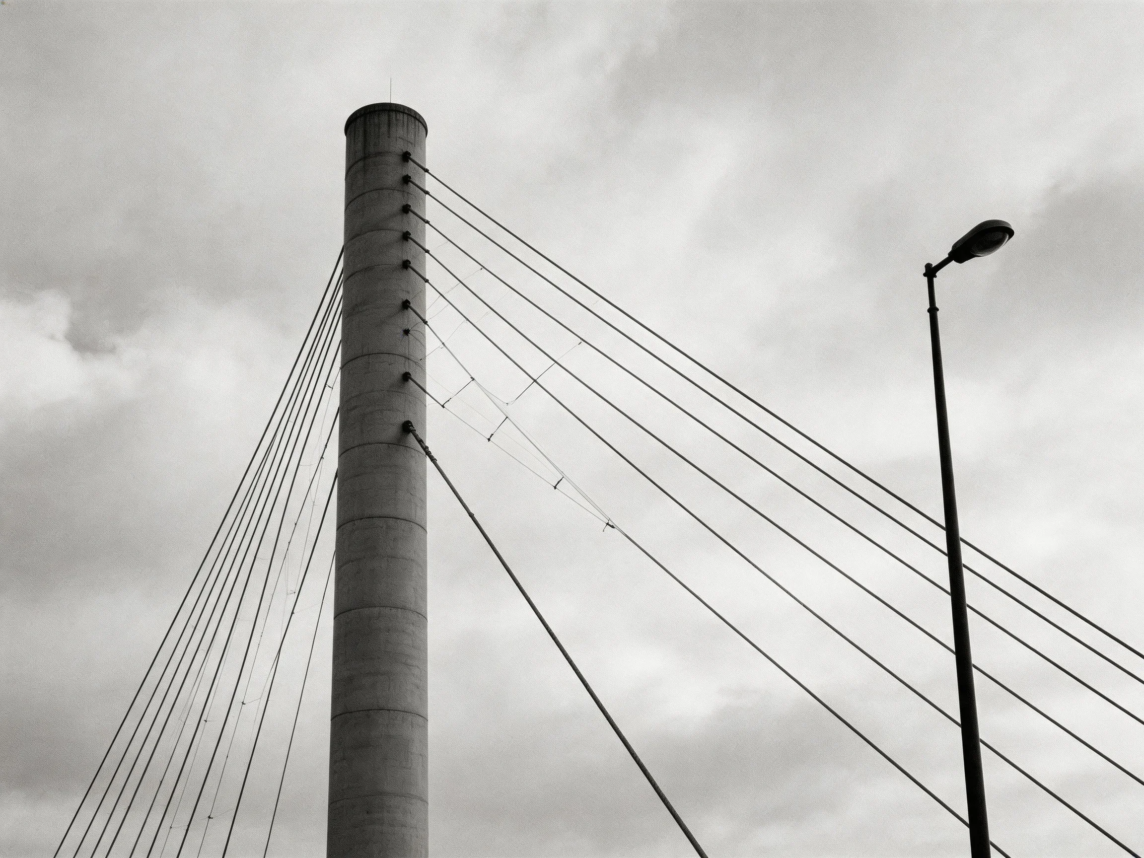 A tall cylindrical tower with angled cables near a streetlight under cloudy sky
