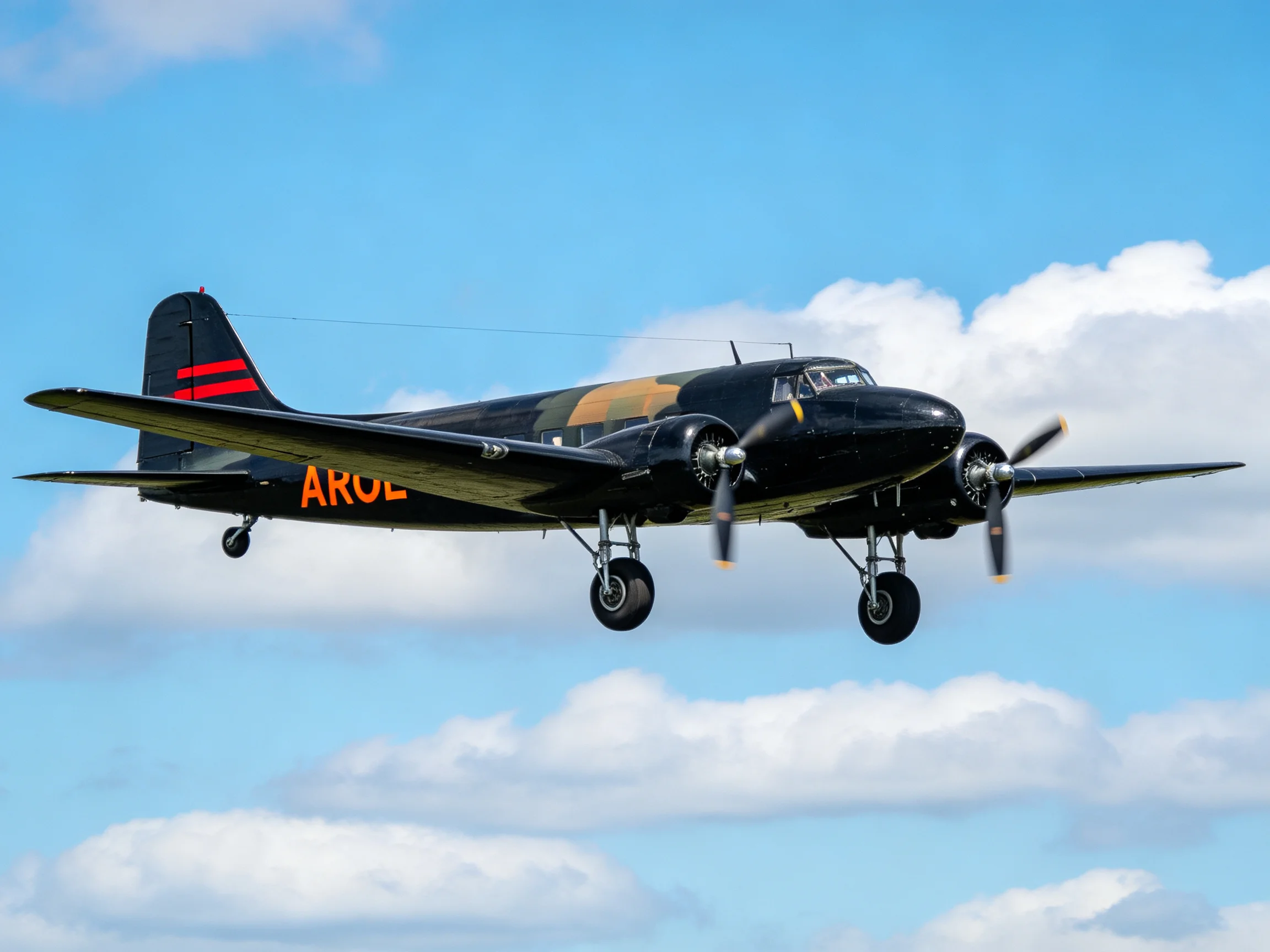 A vintage black twin-engine plane with red tail stripes soars amid fluffy white clouds