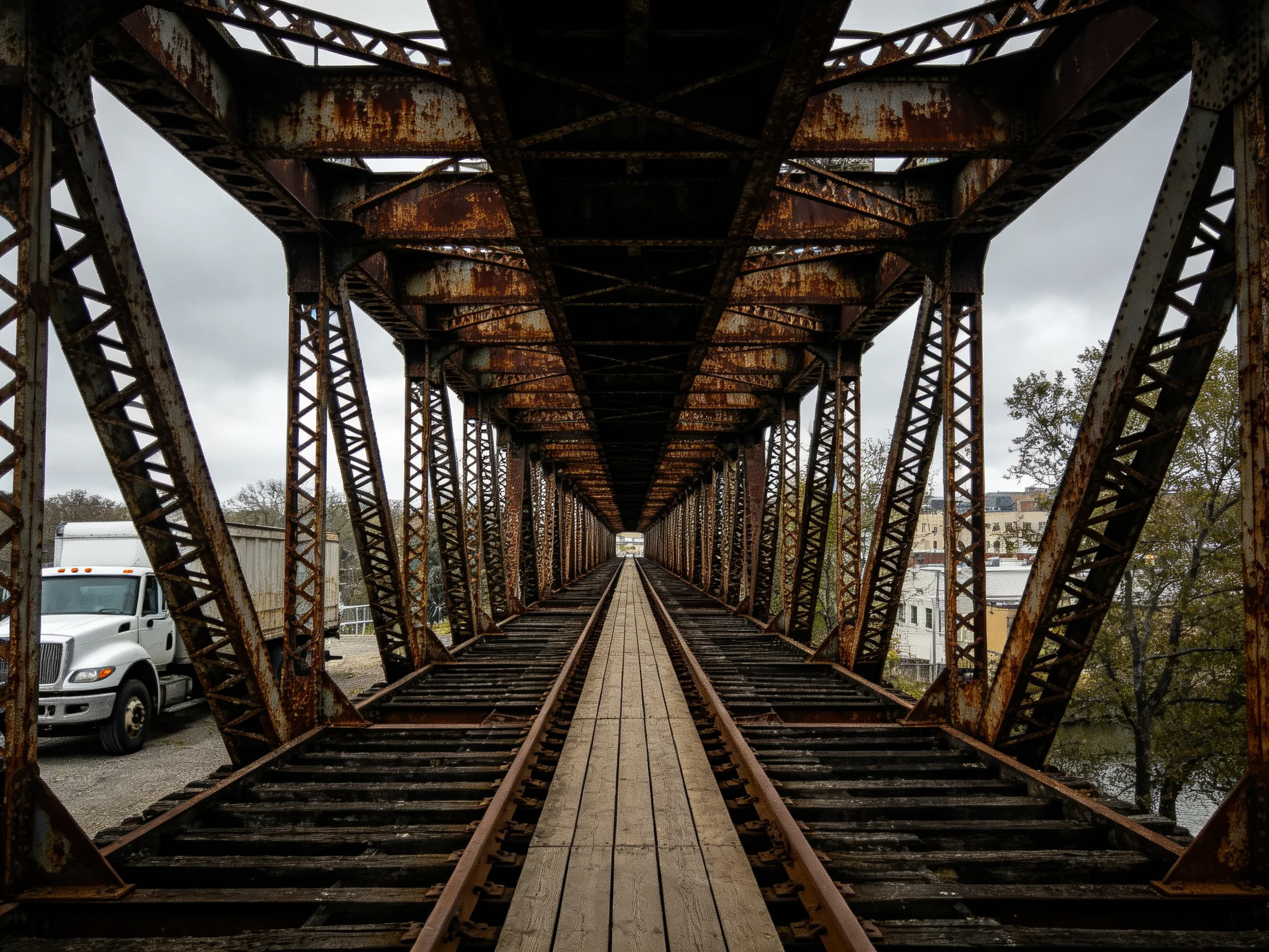 A weathered rust-colored steel bridge spans train tracks under a gray sky