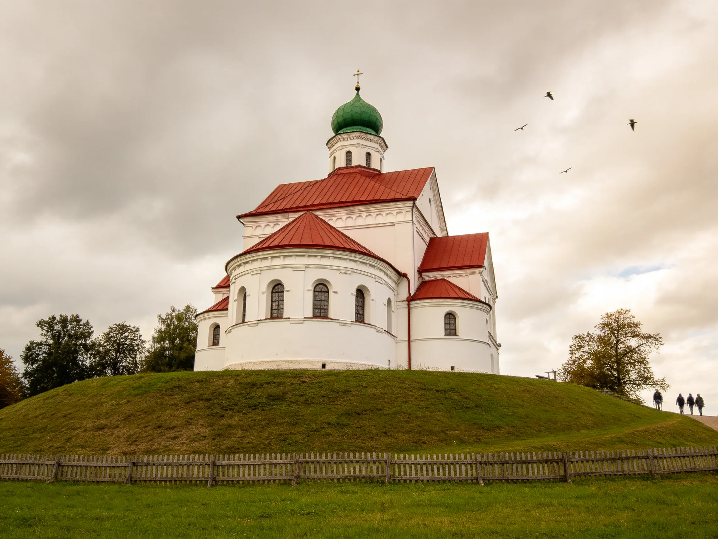 A white church with green dome atop a grassy mound under cloudy sky