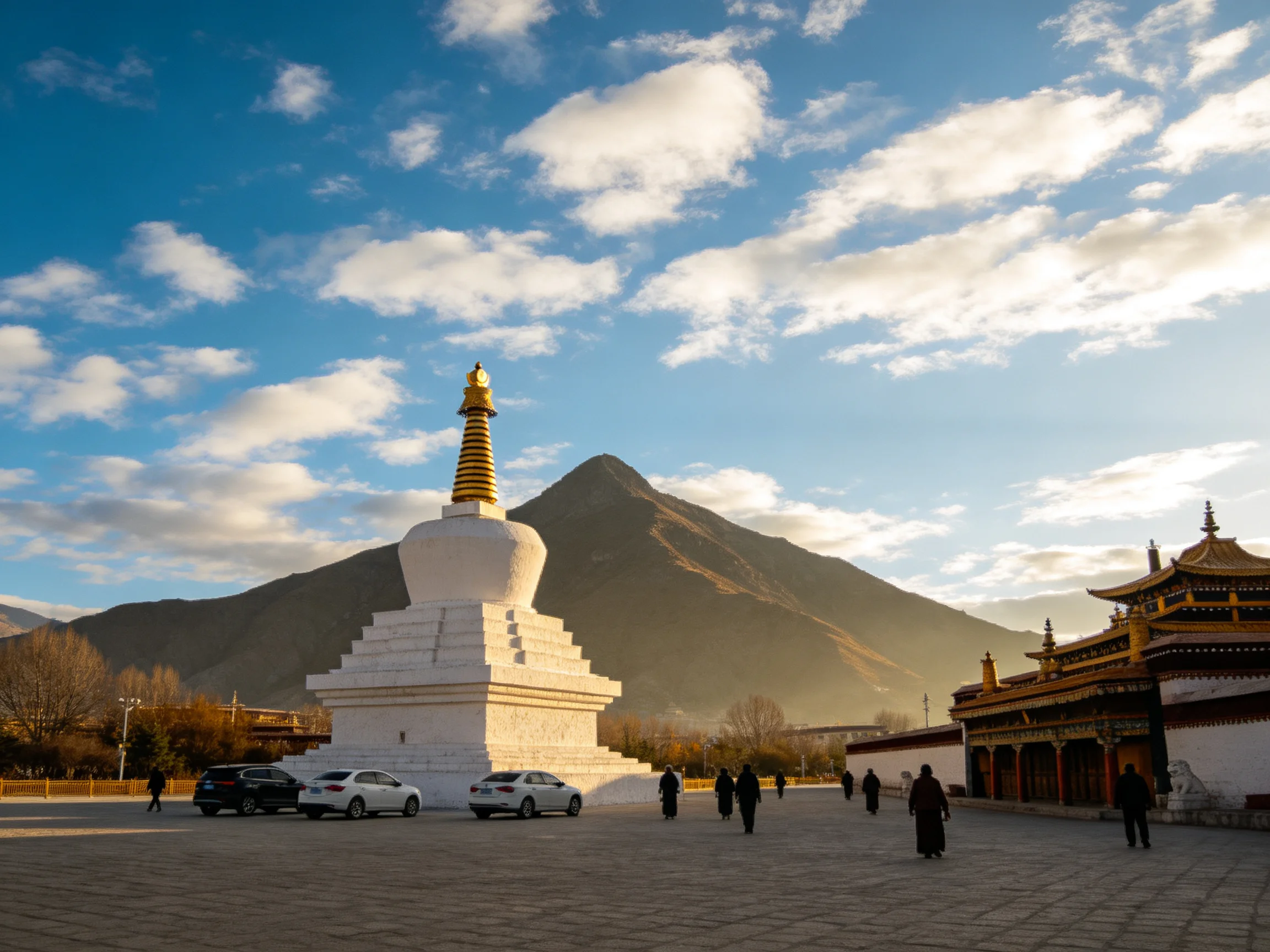 A white stupa and traditional temple are in a plaza with people, near a mountain.