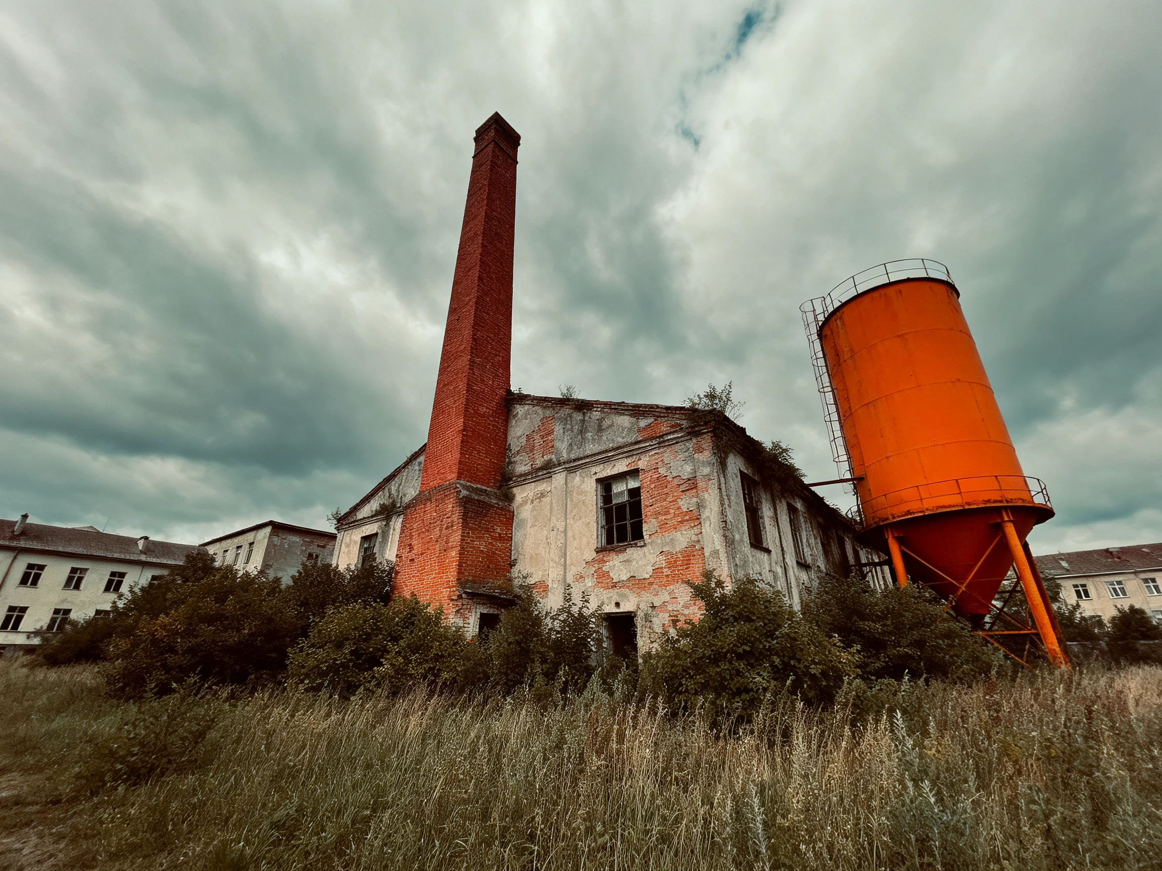 An abandoned brick factory with a tall red chimney and bright orange silo