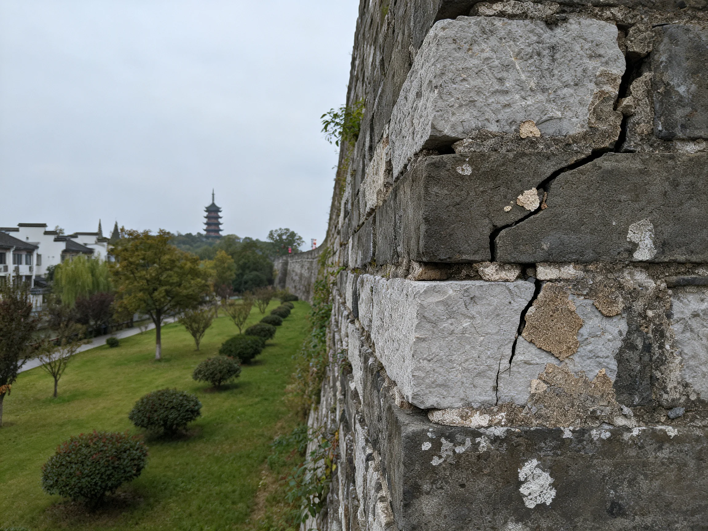 Ancient stone wall with cracked gray stones near green park and white buildings