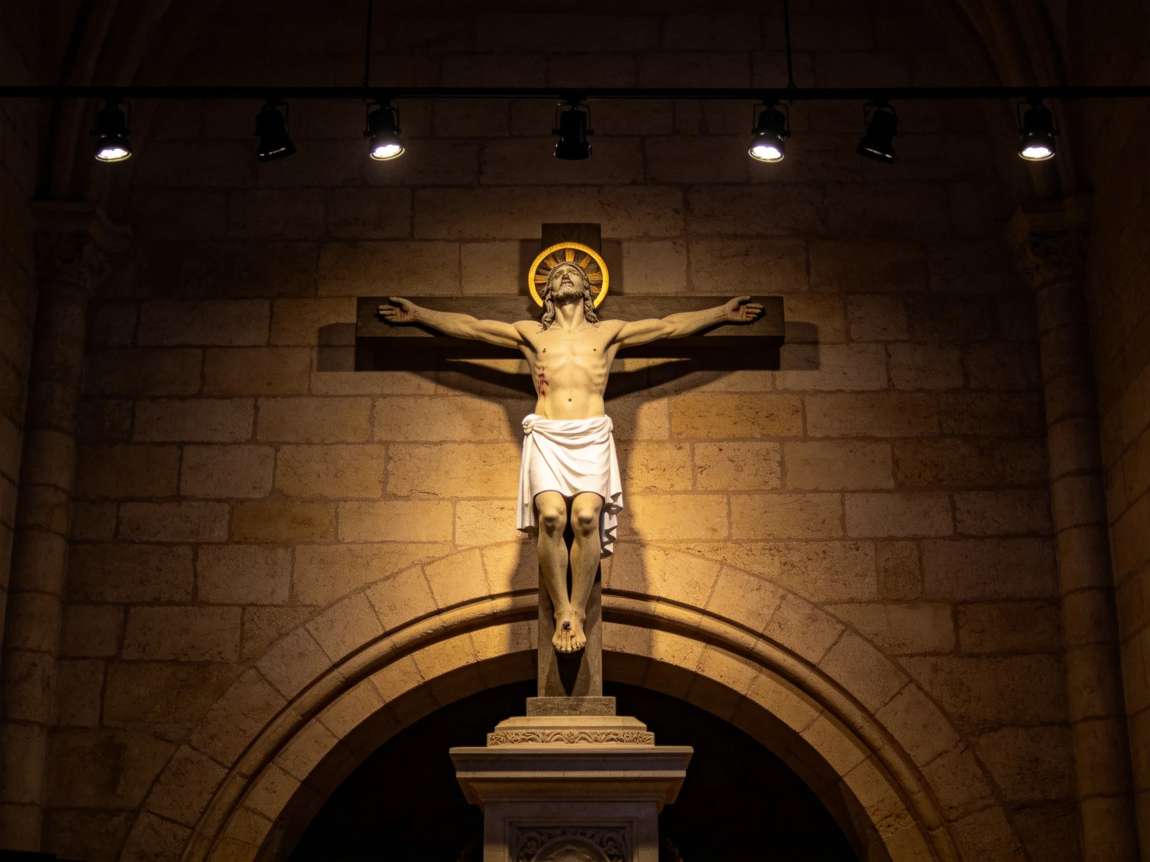 Crucifix sculpture with golden halo on stone wall under spotlights