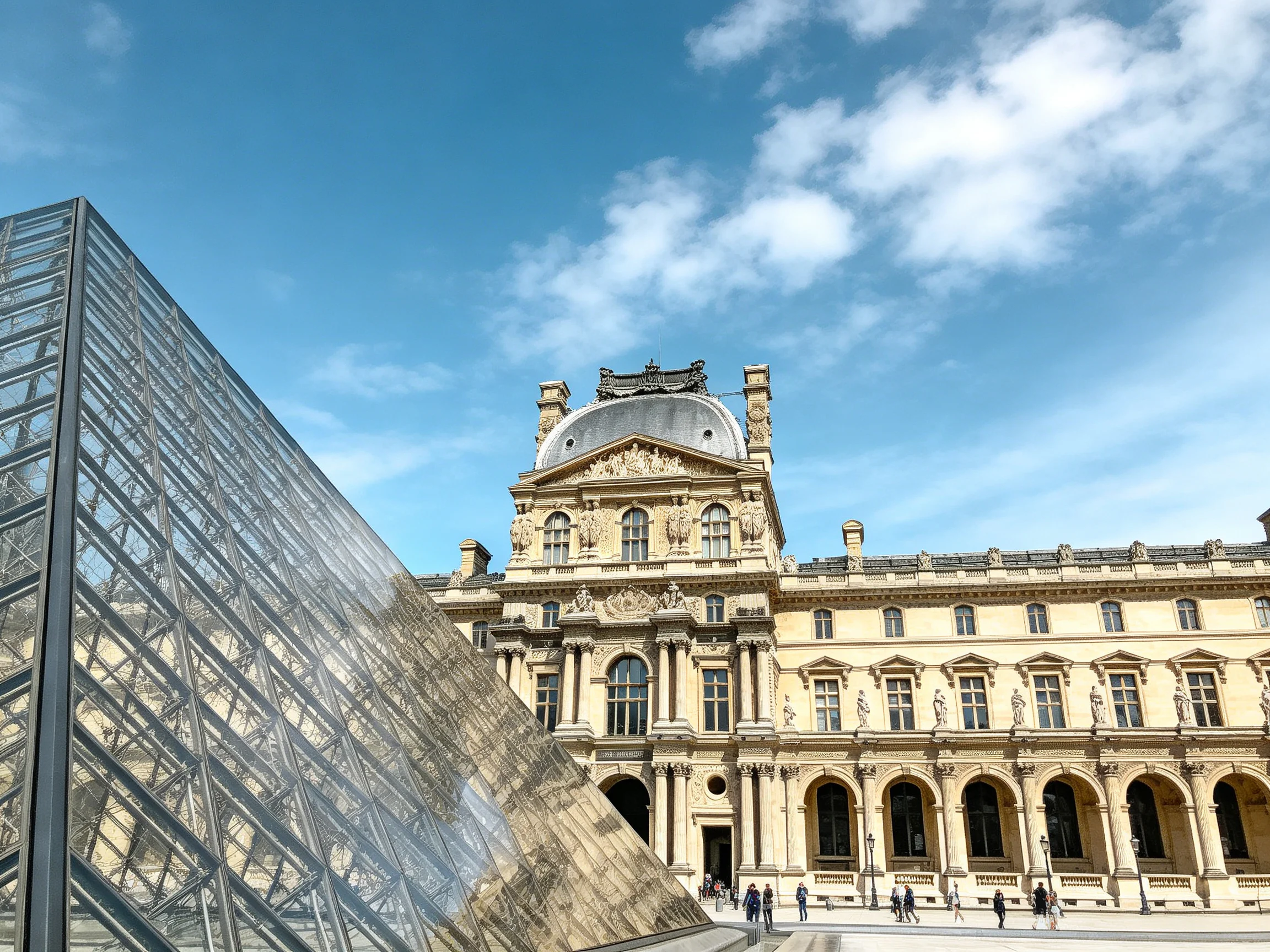 Modern glass pyramid beside ornate historical museum under clear blue sky