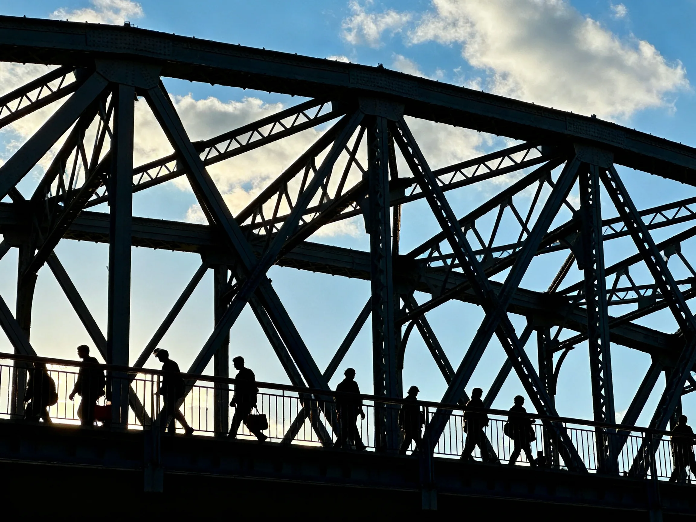 Silhouetted pedestrians traverse a large iron truss bridge under a bright sky