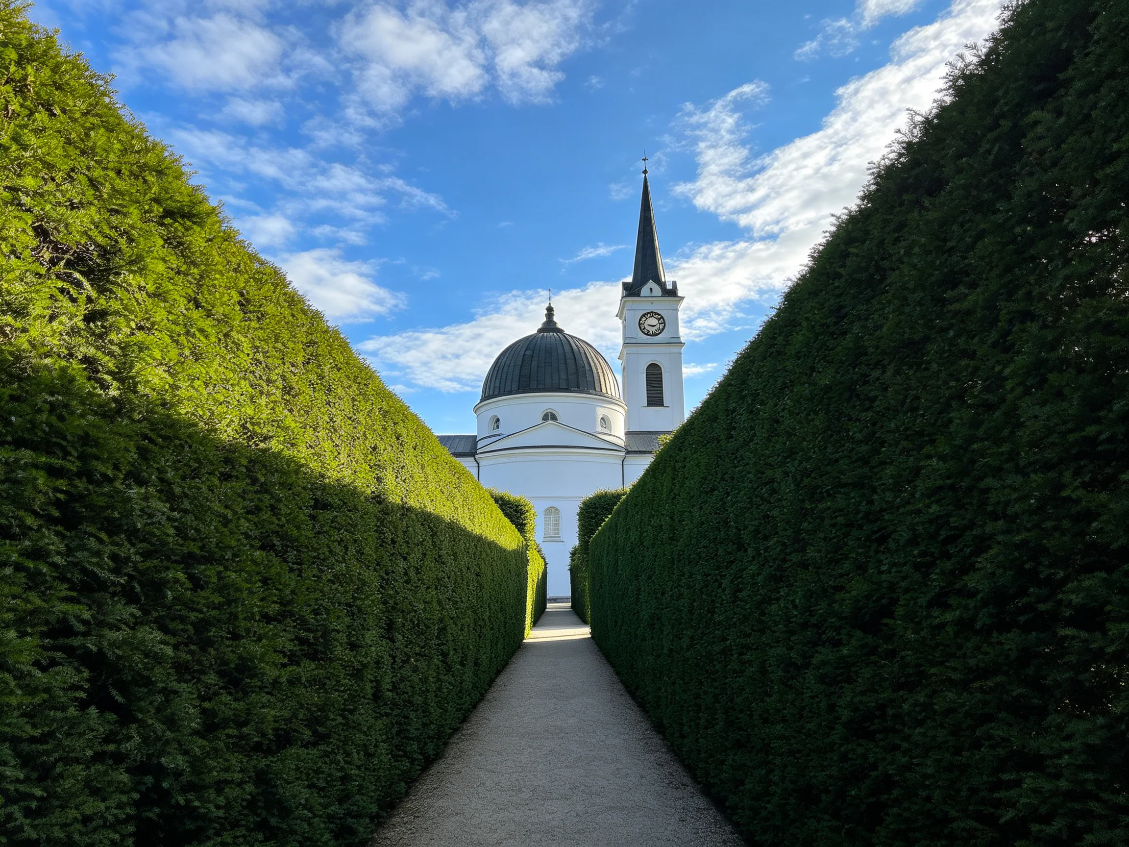 Tall green hedges frame a white domed church with clock tower
