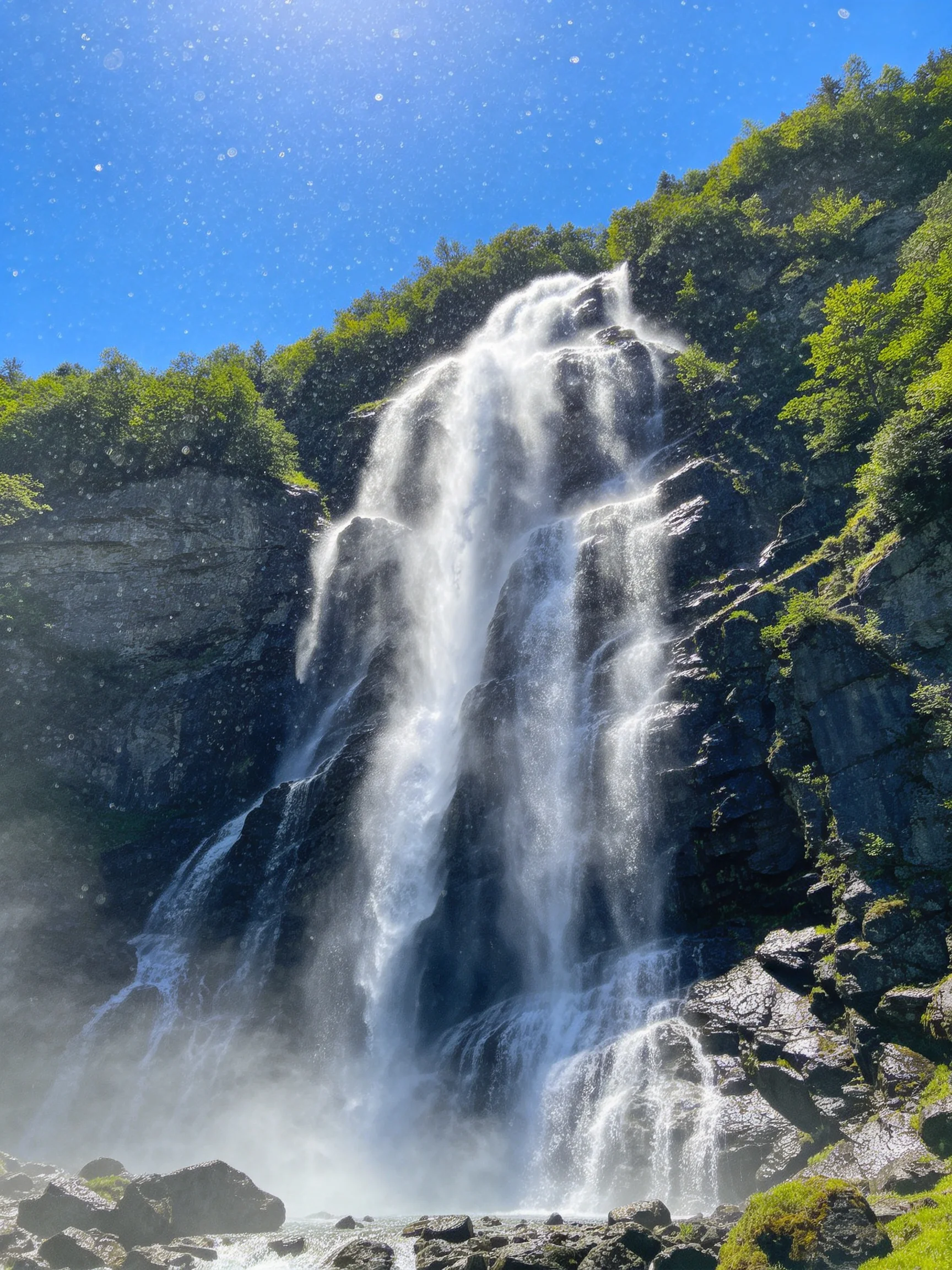 A powerful waterfall cascades over dark rocky cliffs amid lush green foliage