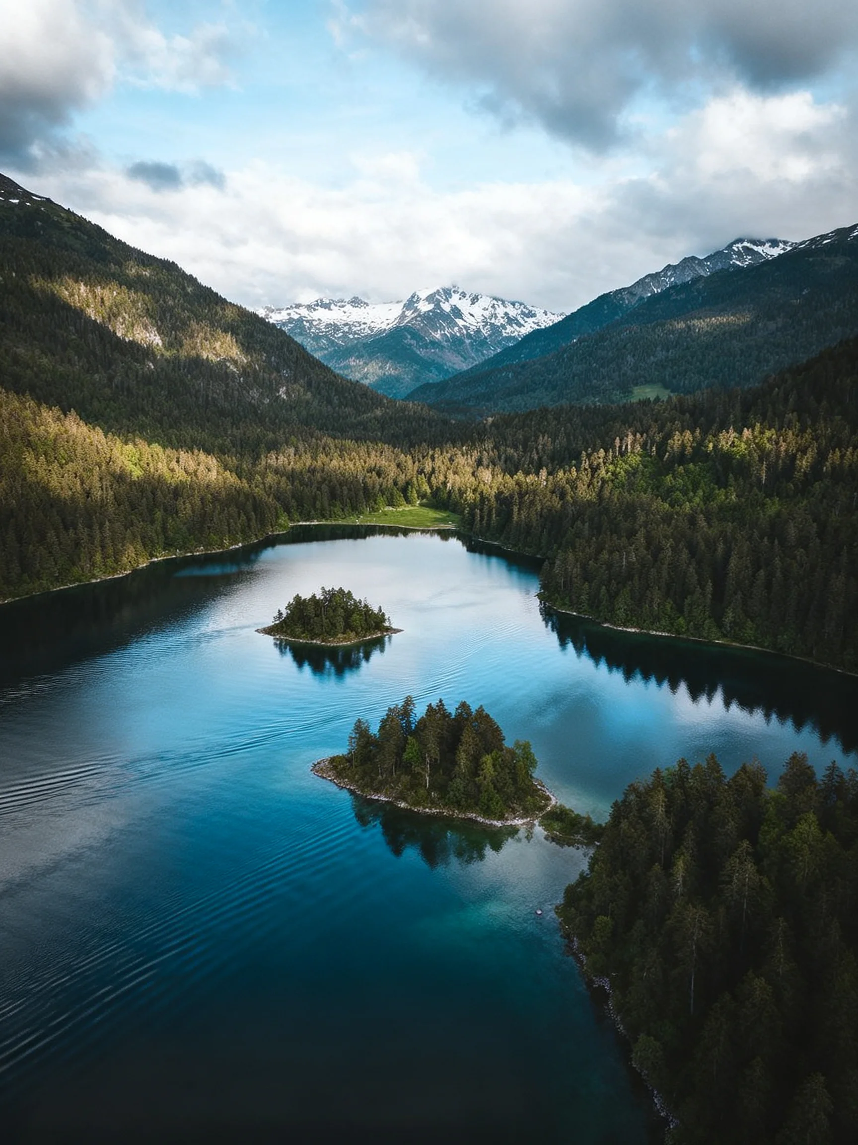 A serene lake with two forested islands, surrounded by dense green forests and snow-capped mountains.