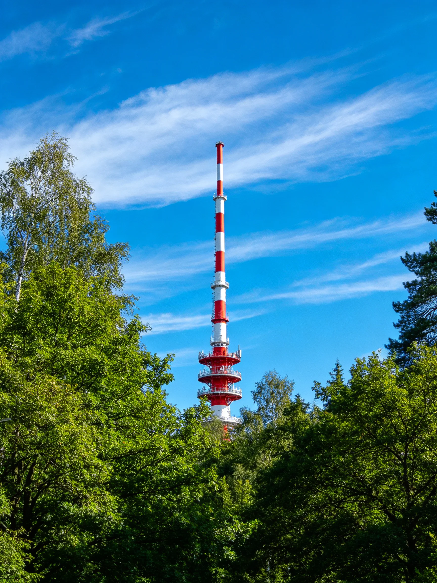 A tall red-and-white tower rises amid lush green trees under bright blue sky