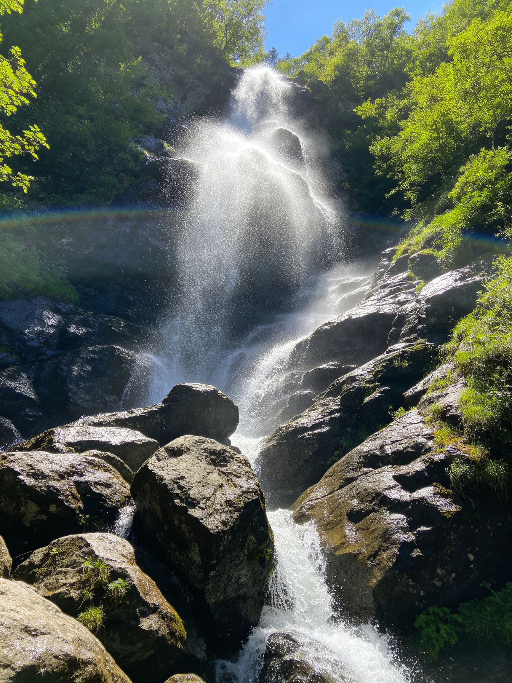 A waterfall with misty spray flows over moss-covered dark rocks near green trees