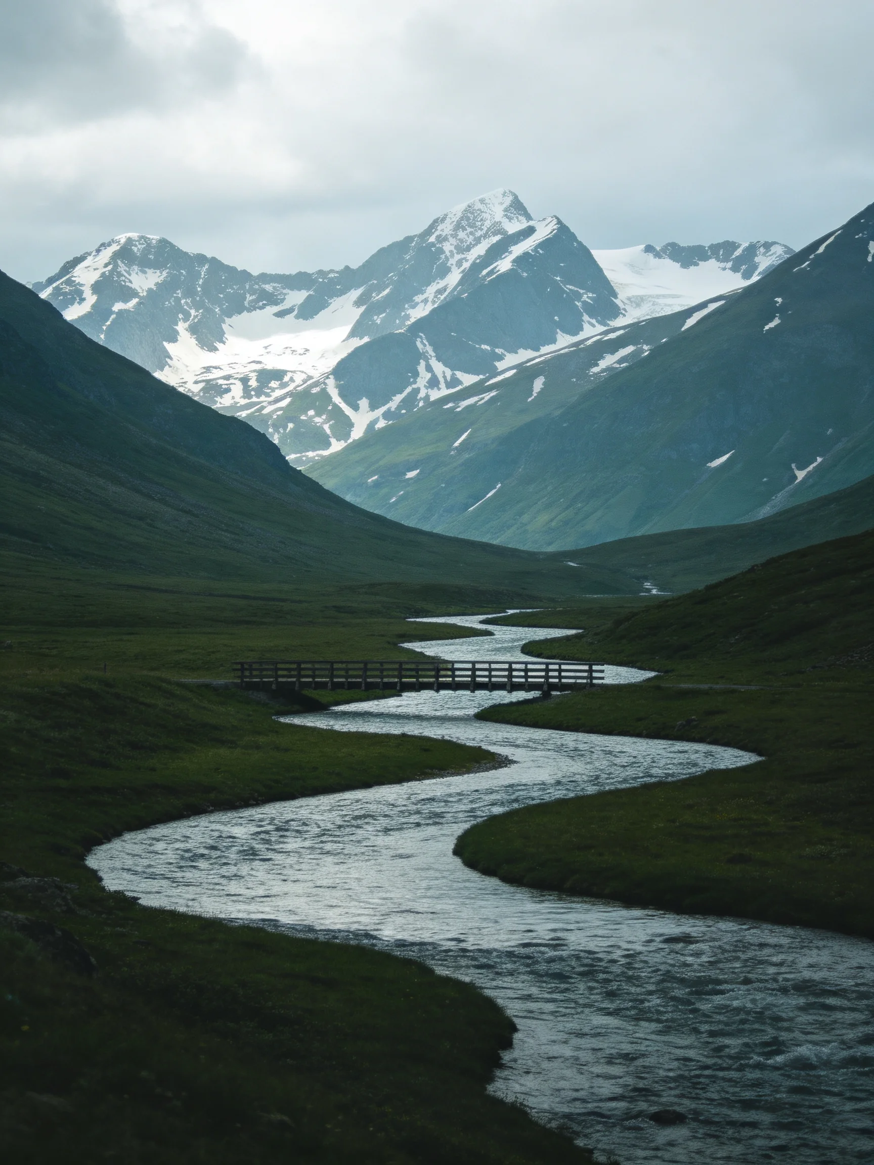Snow-capped peaks tower over a winding river with a wooden bridge