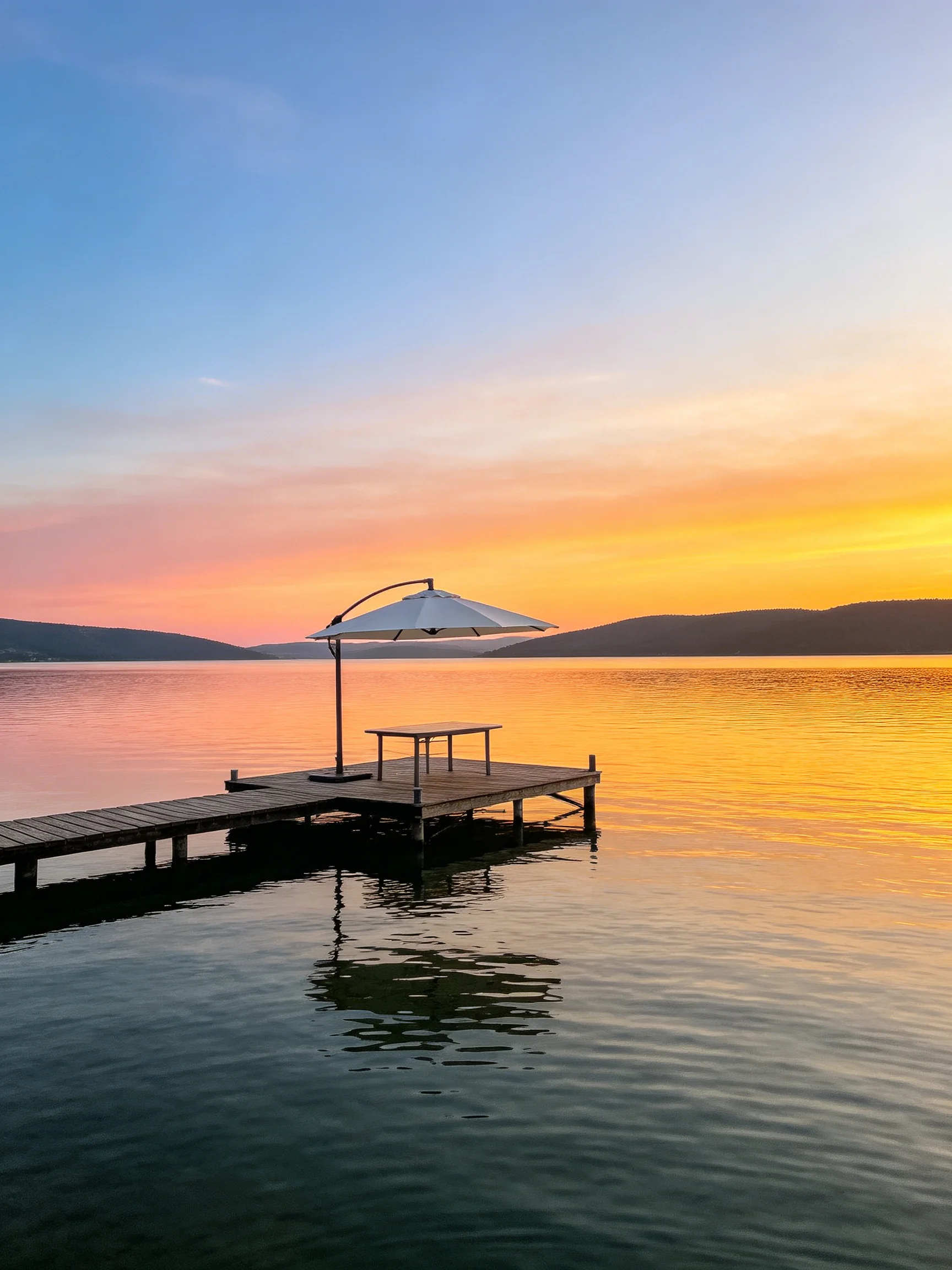 Wooden dock with umbrella overlooks calm lake at colorful sunset