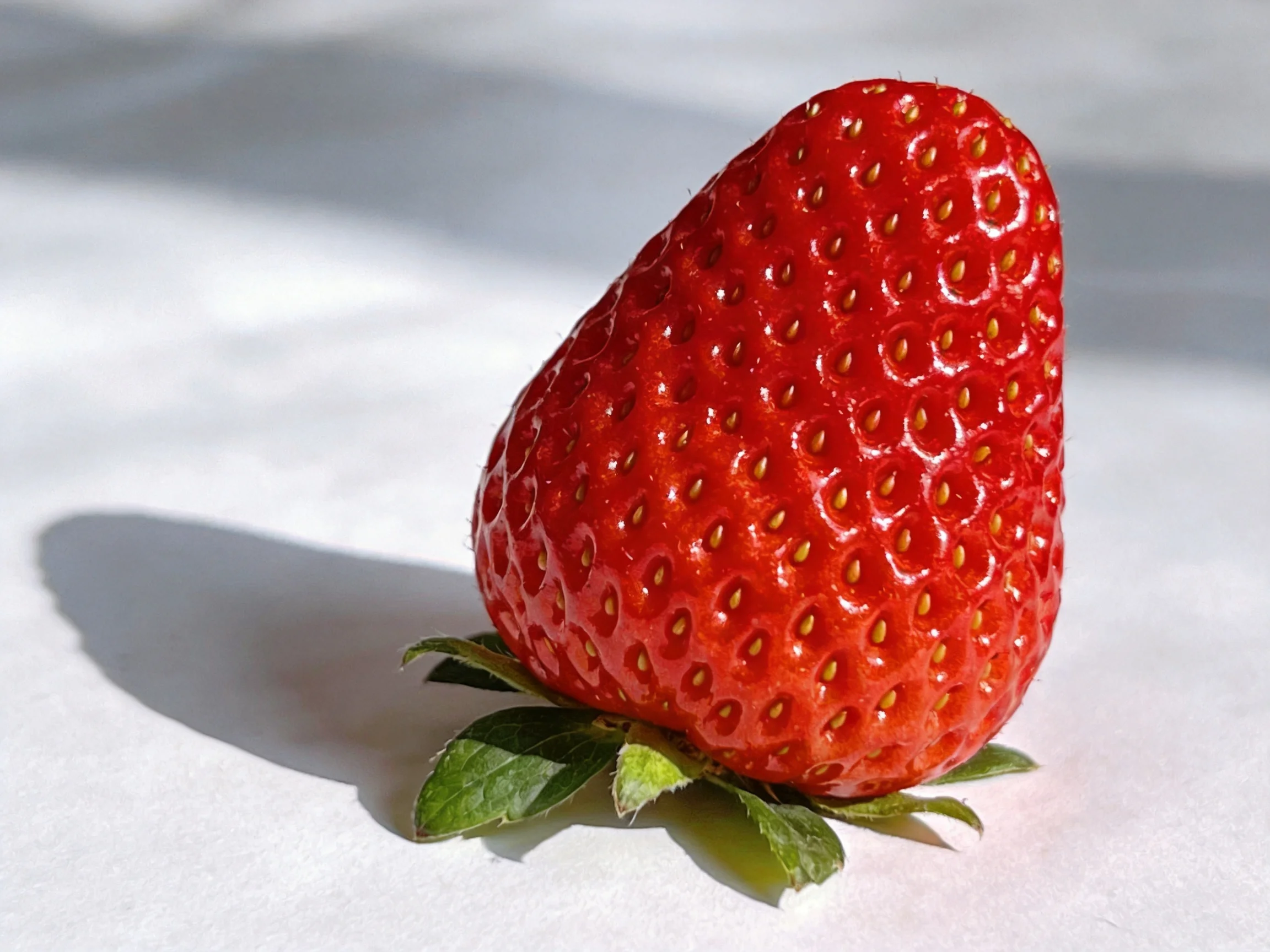 A bright red strawberry with green foliage rests on a white surface