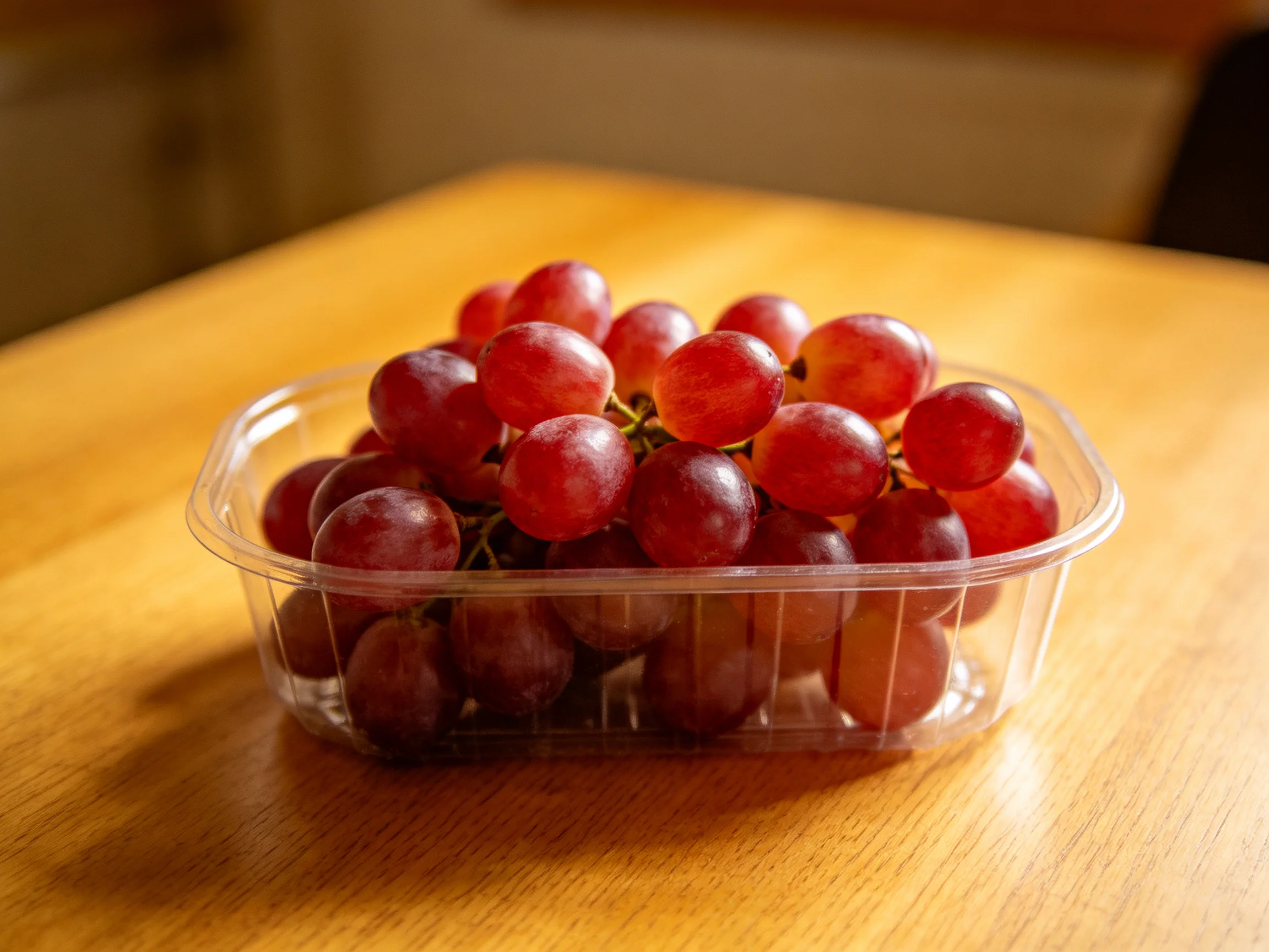 A clear plastic container holds vibrant red grapes on a wooden table
