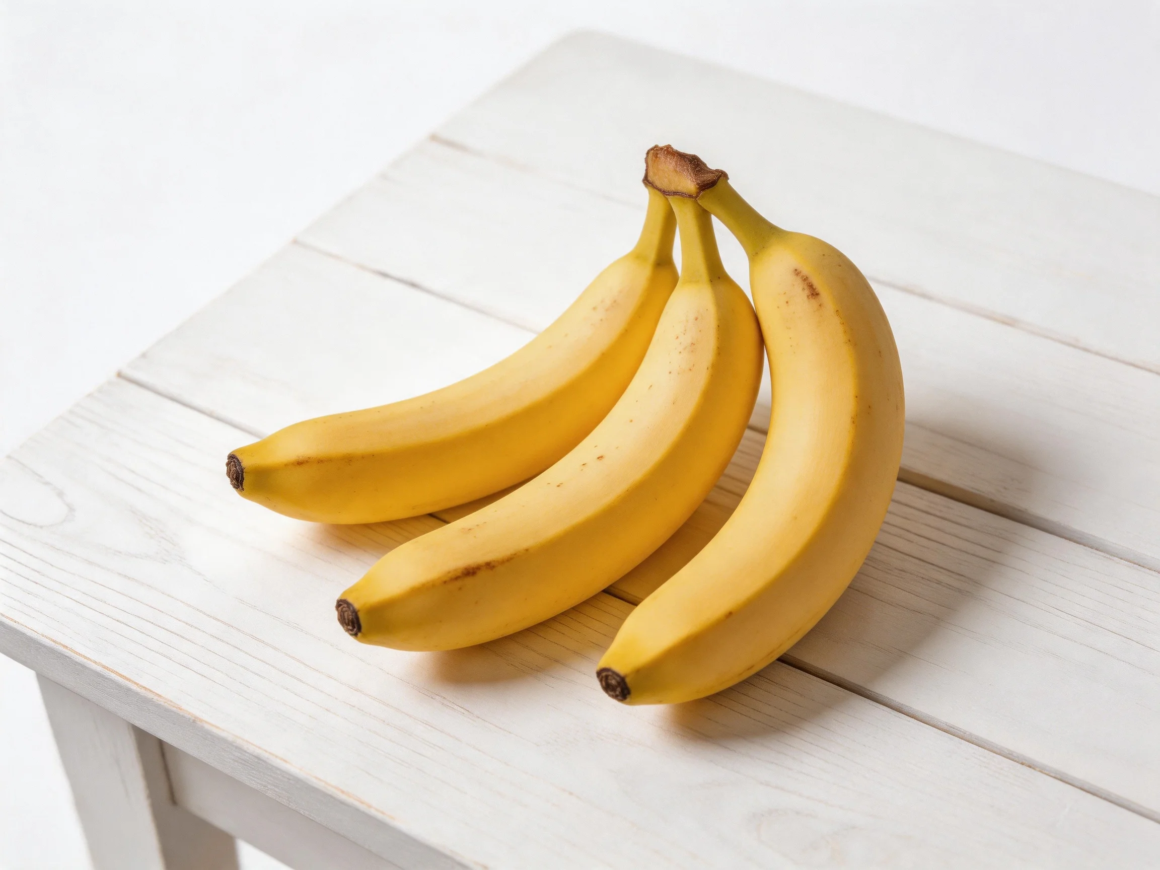 A cluster of three ripe yellow bananas sits on a white wooden table.