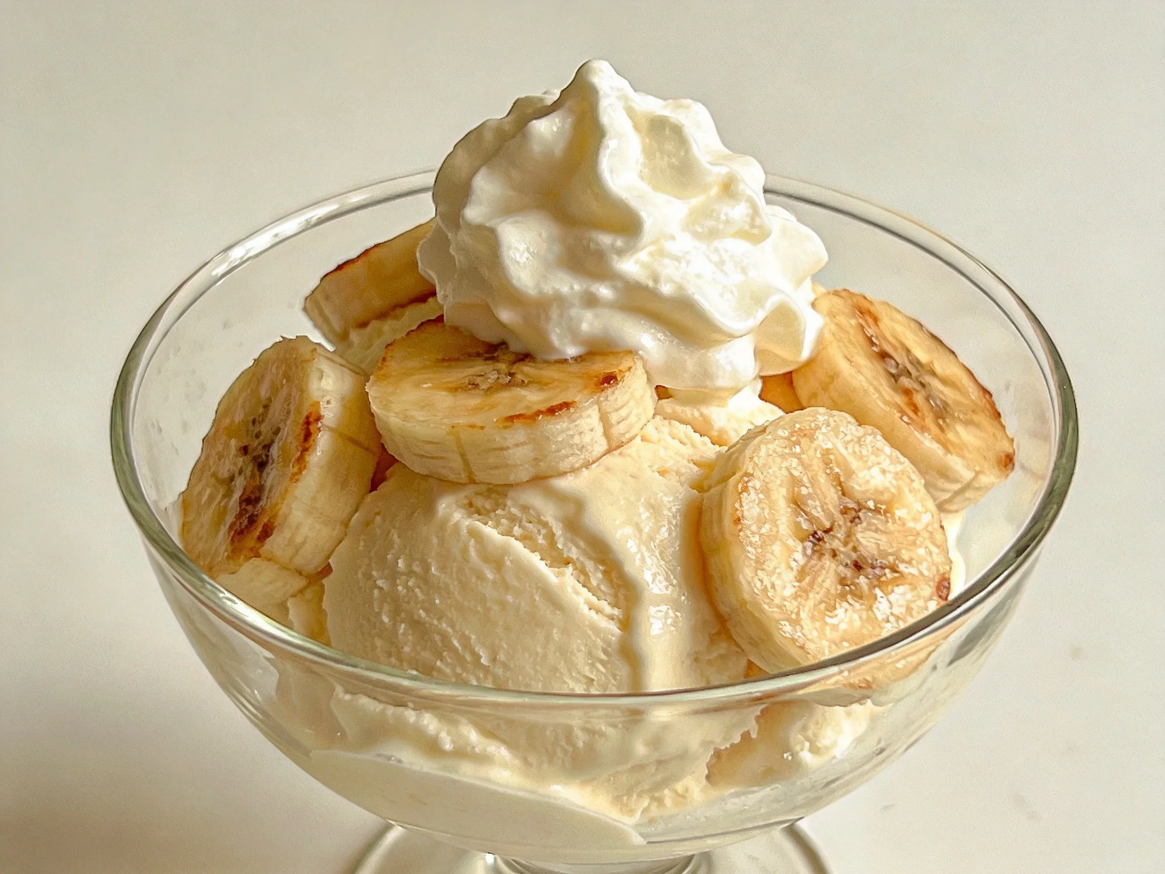 A glass dessert dish holds vanilla ice cream, caramelized banana slices, and whipped cream.