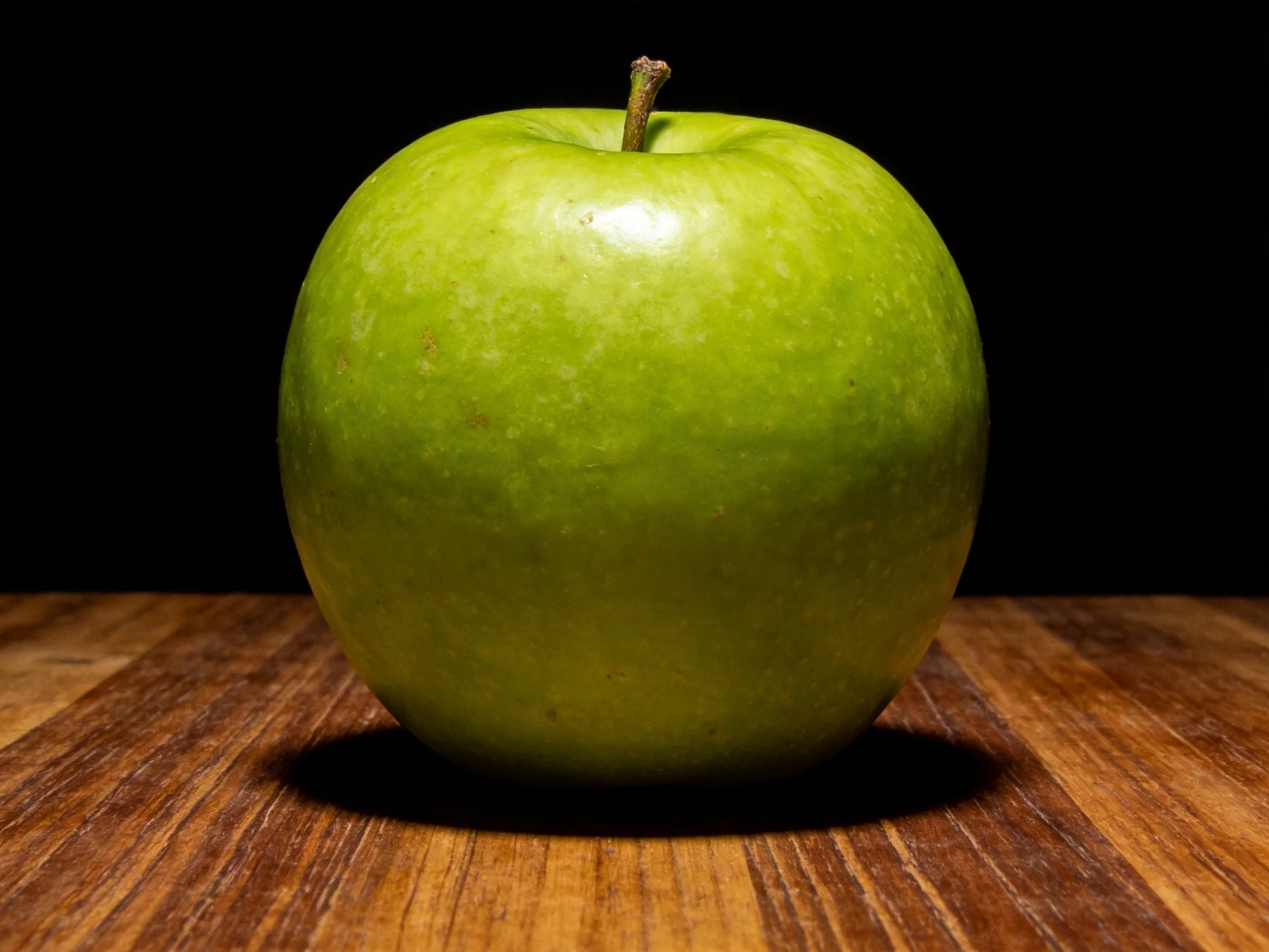 A single bright green apple sits on a wooden surface with a dark backdrop