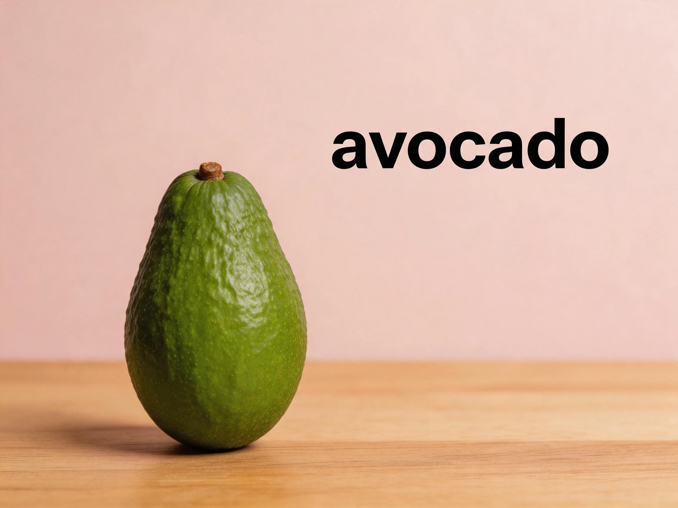 A single green avocado sits on a light wooden surface with a pink backdrop