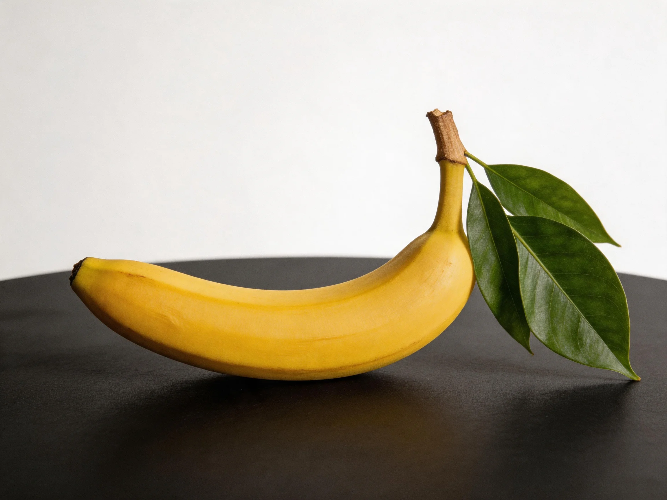 A single ripe yellow banana with green leaves rests on a dark surface