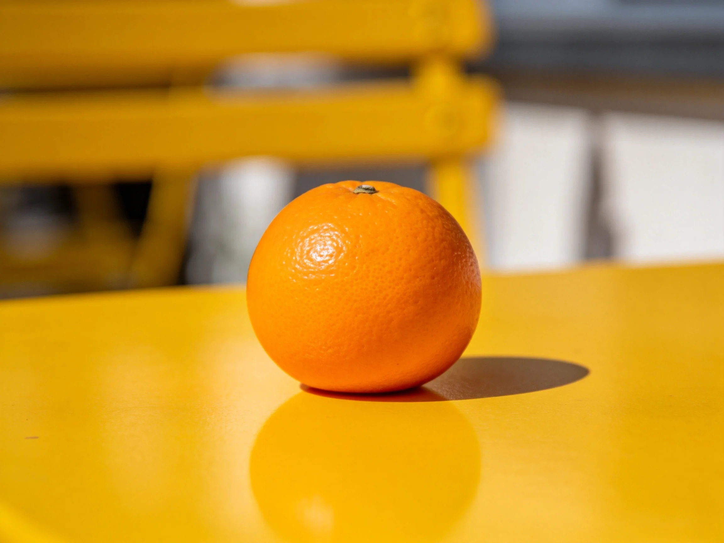 A single vibrant orange rests on a glossy yellow table beside a yellow bench