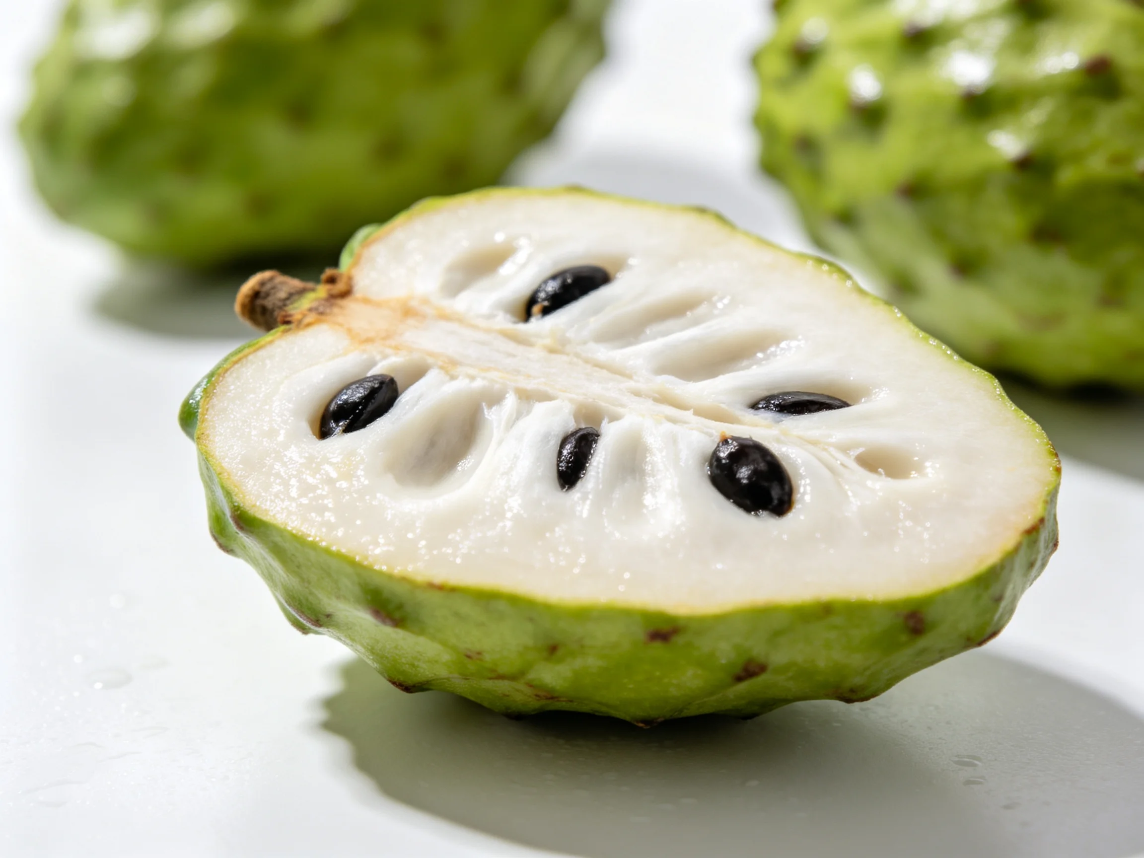 A sliced soursop fruit shows green exterior and white flesh with black seeds