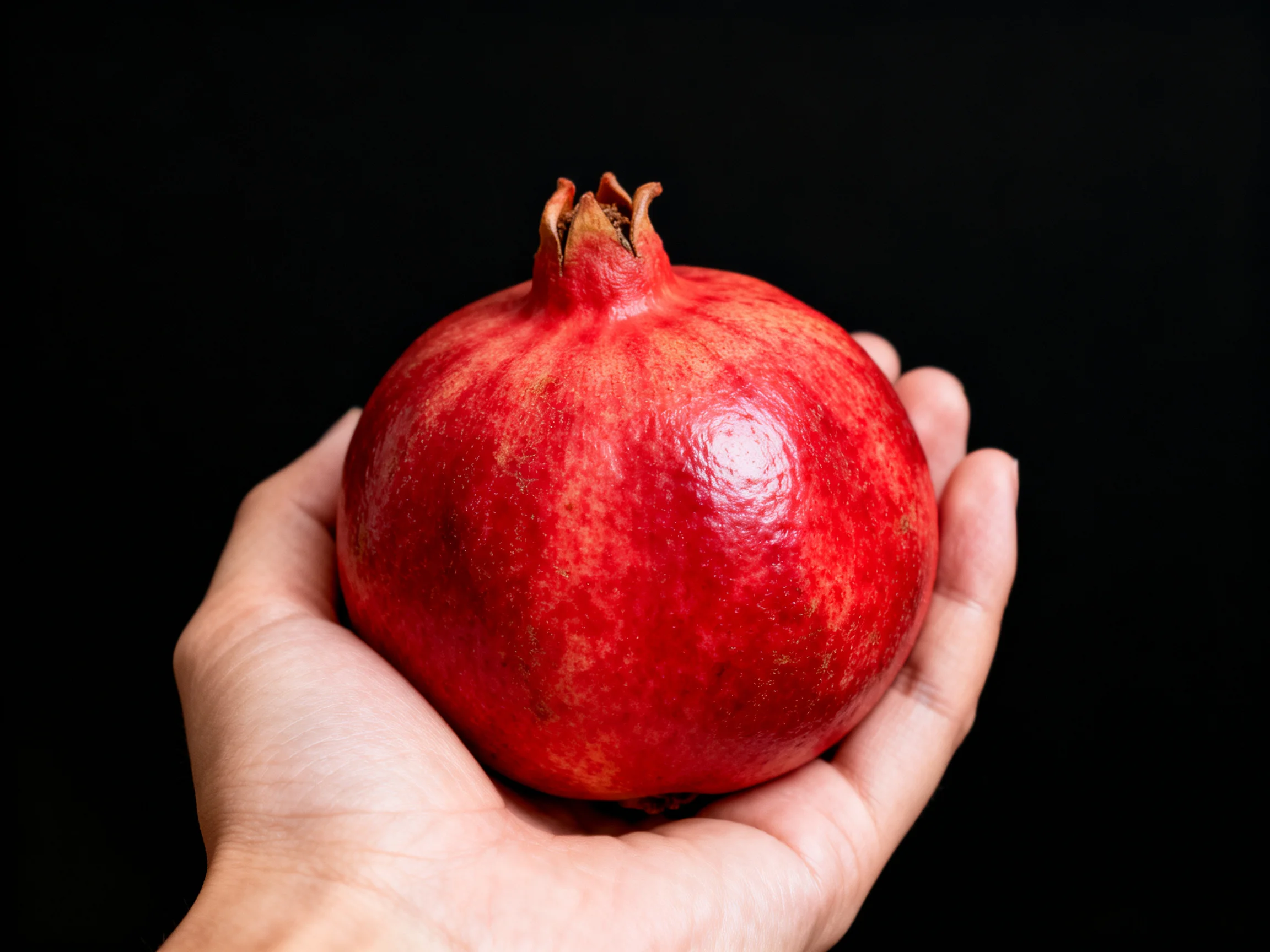 A vibrant red pomegranate cradled in an open hand with a black backdrop