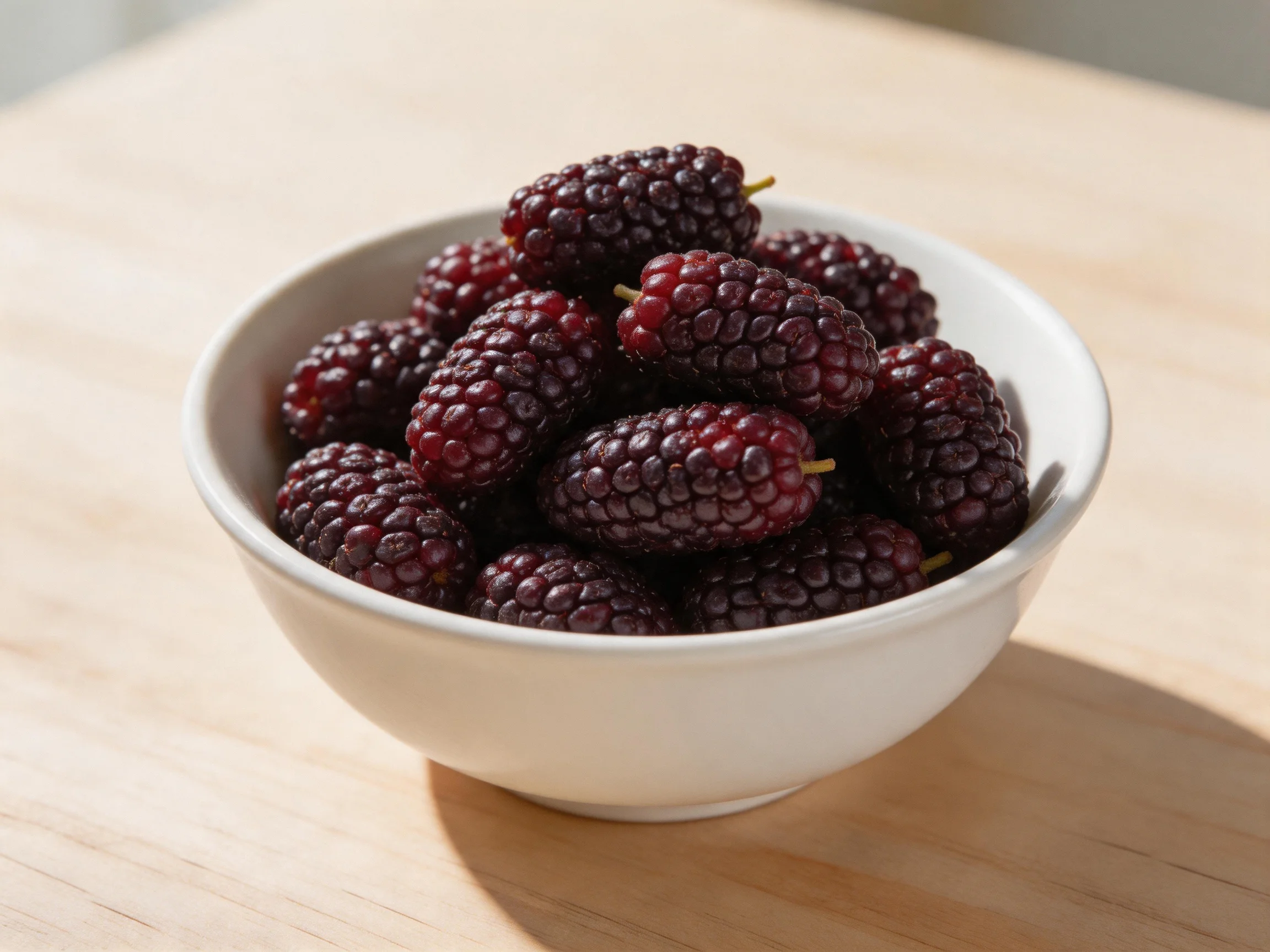 A white bowl brimming with deep purple mulberries on a wooden surface