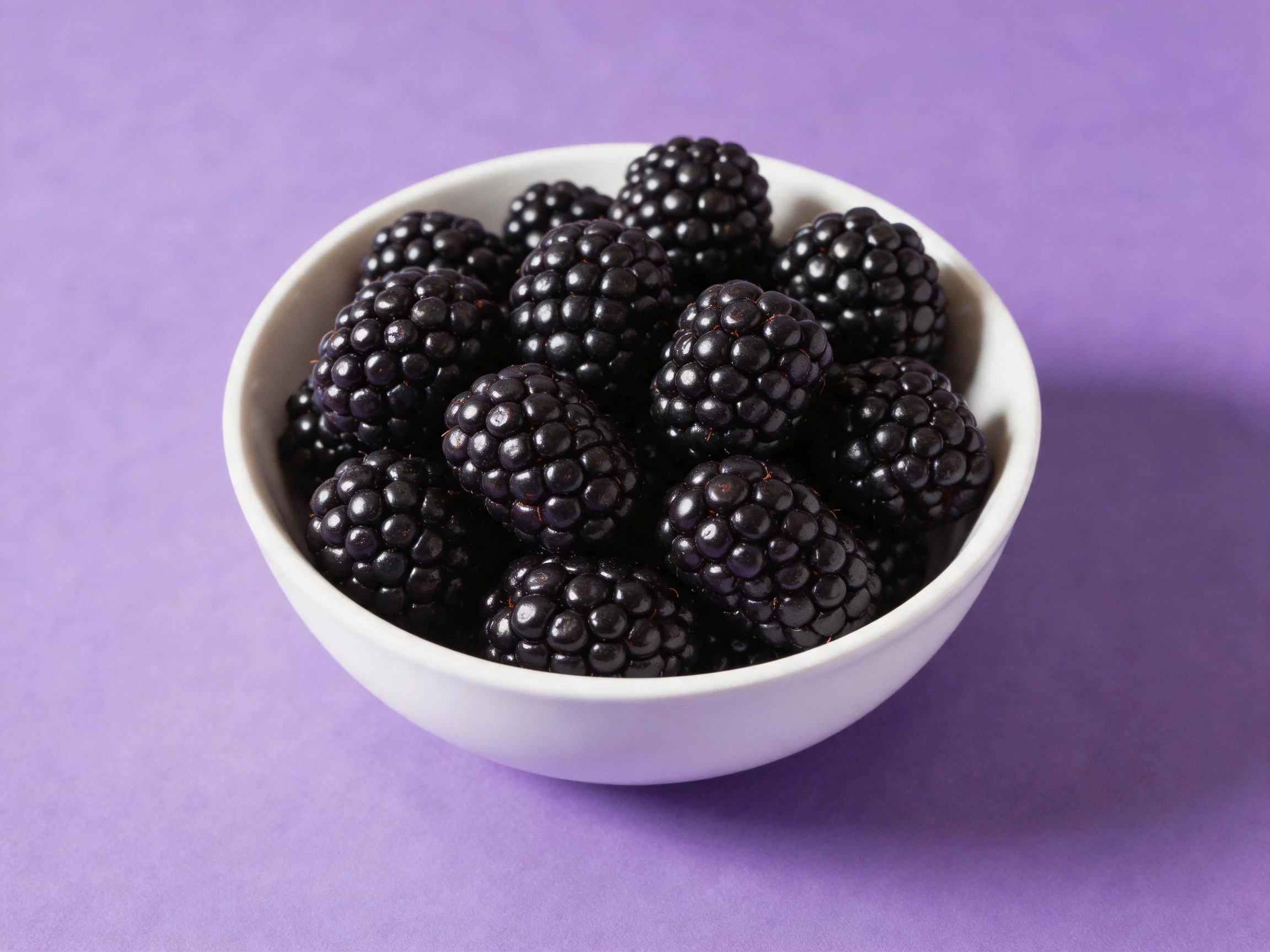 A white bowl filled with glossy blackberries on a vibrant purple surface