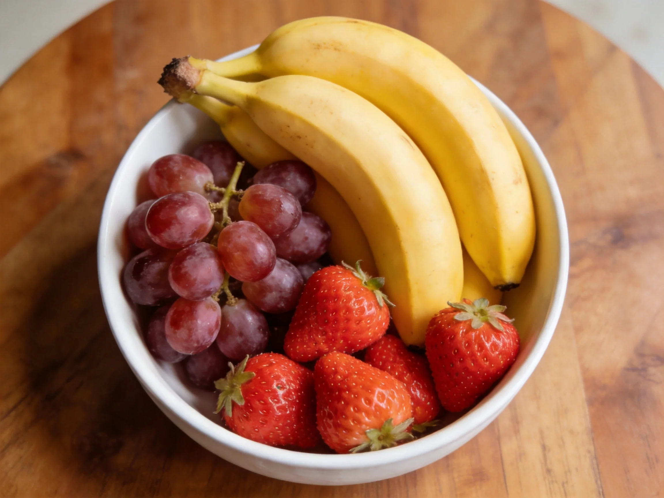 A white bowl holds yellow bananas, purple grapes, and red strawberries.