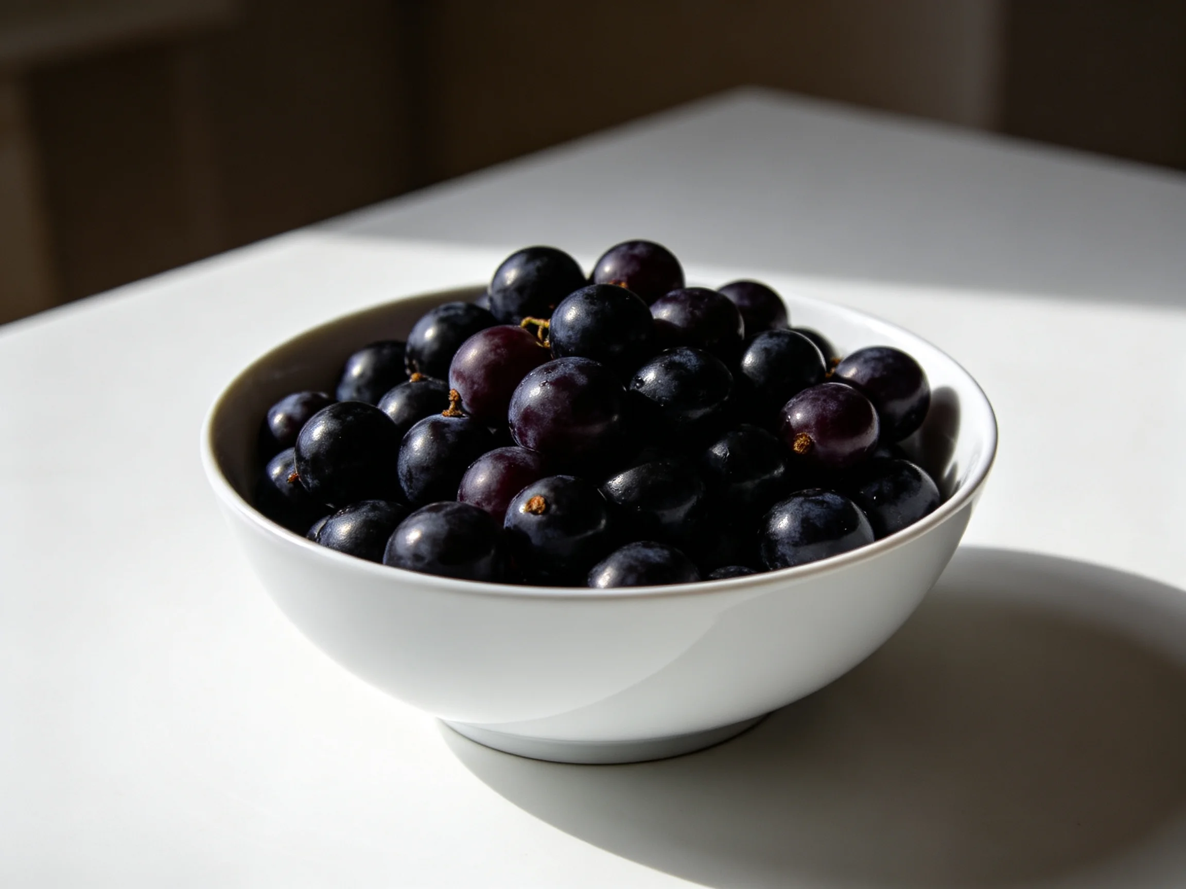 A white bowl overflowing with glossy dark purple grapes on a bright surface