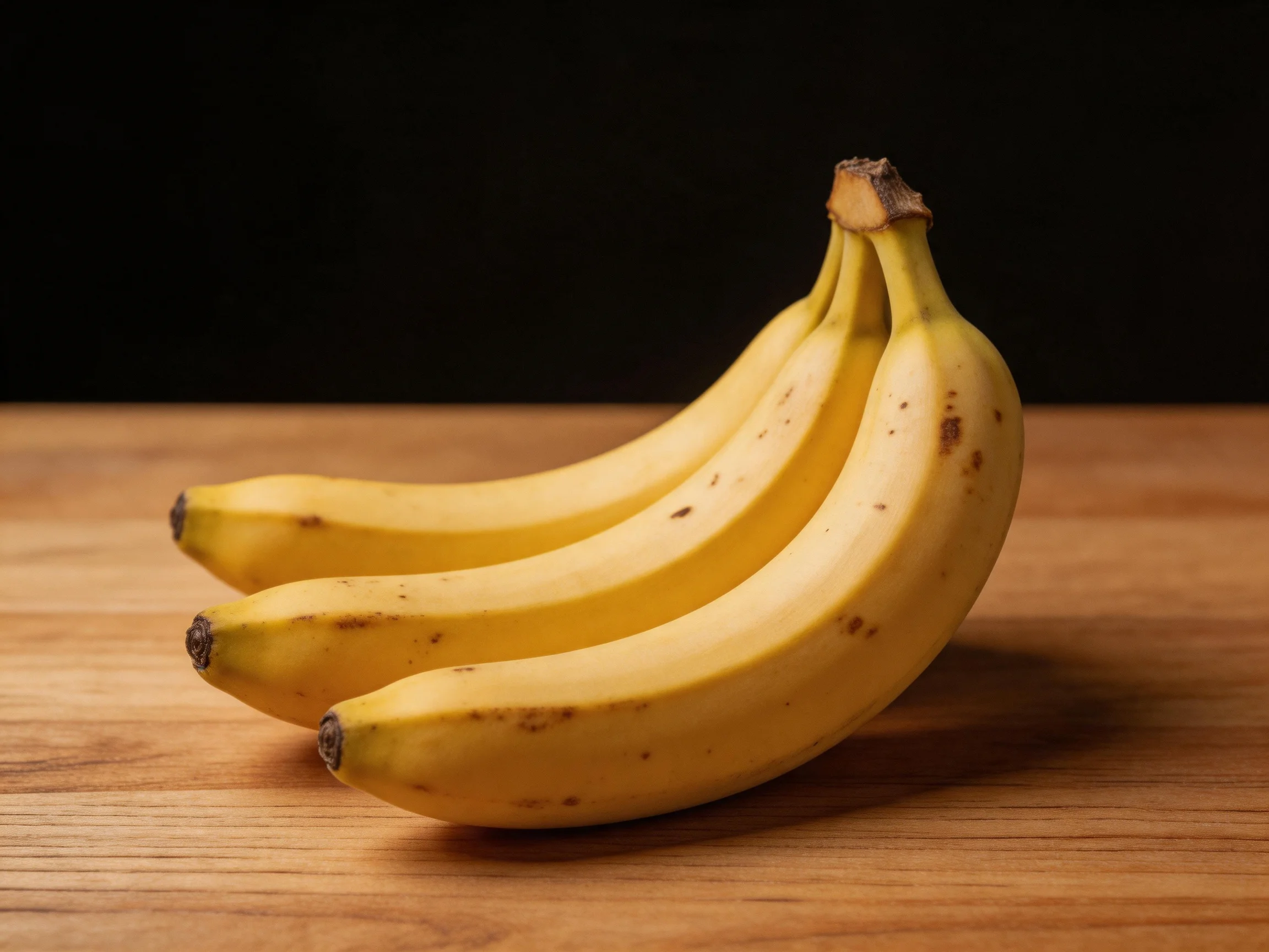 Three ripe yellow bananas arranged on a wooden surface with a dark backdrop