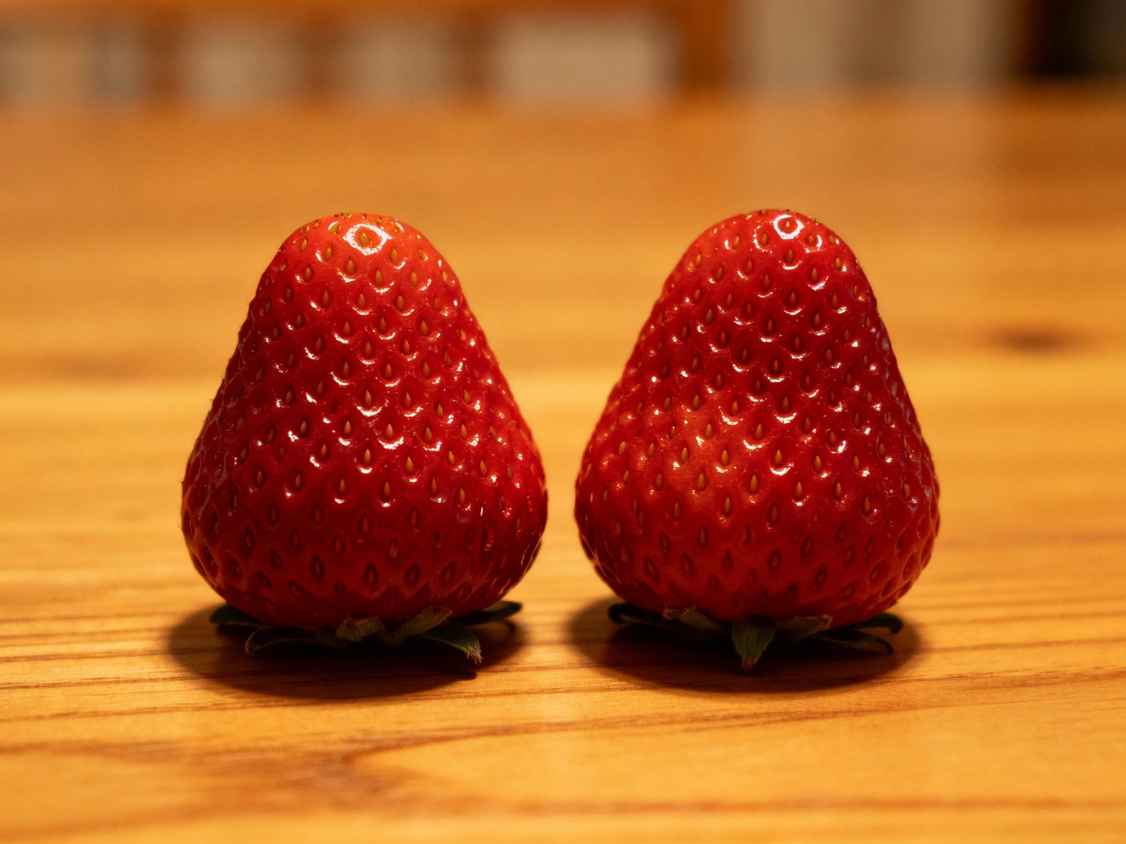 Two glossy red strawberries positioned side by side on a wooden table