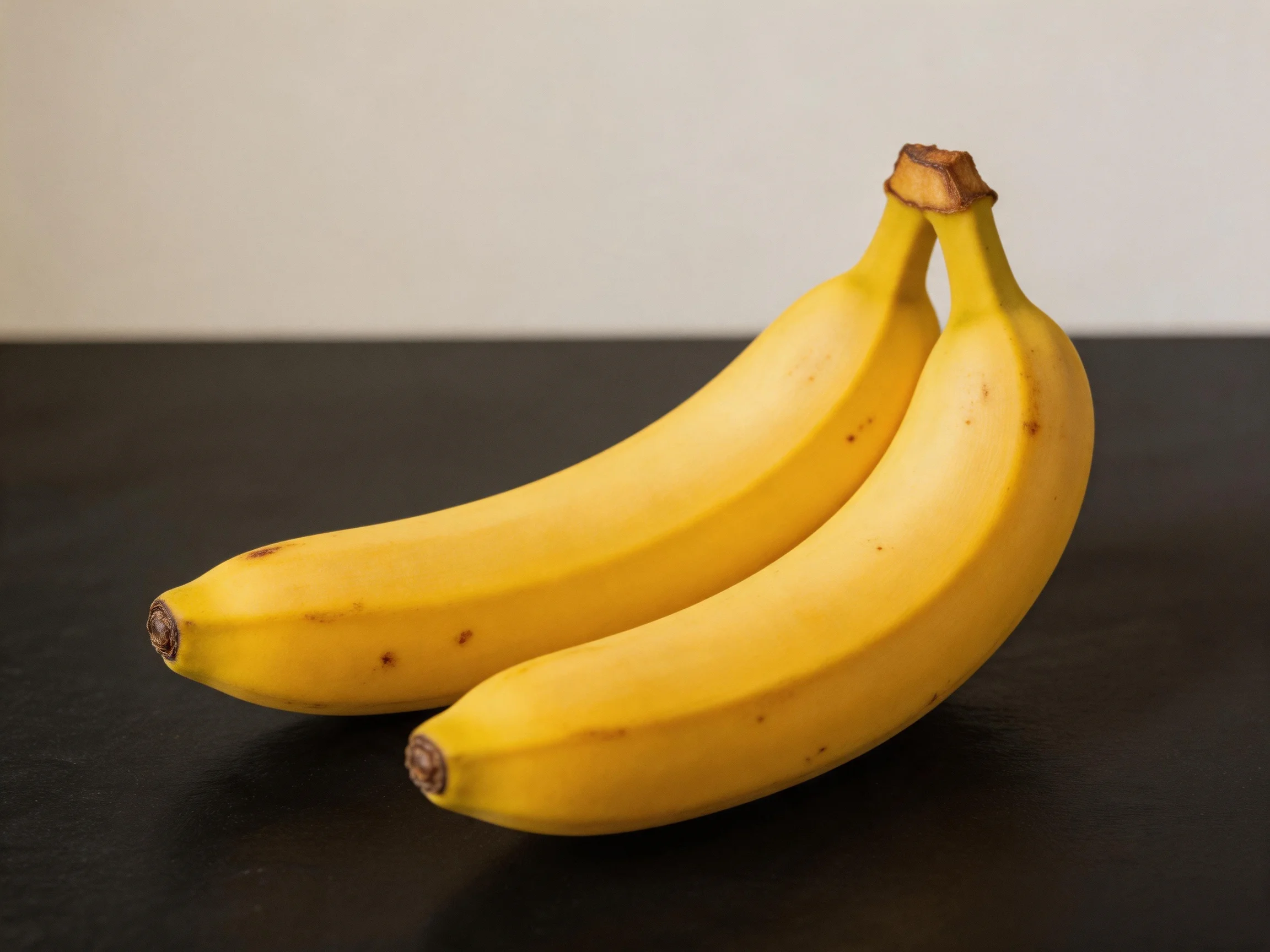 Two ripe yellow bananas on a dark surface with a plain wall