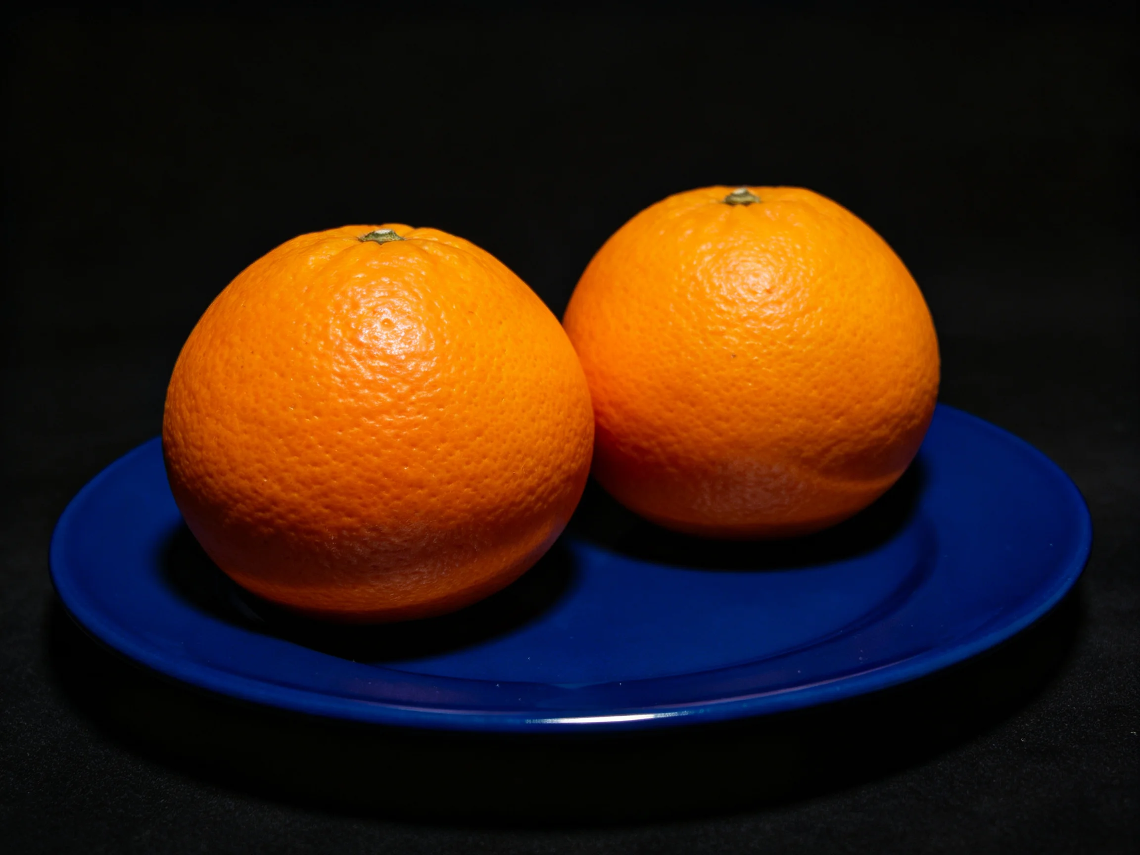 Two vibrant oranges rest on a deep blue plate with a dark backdrop