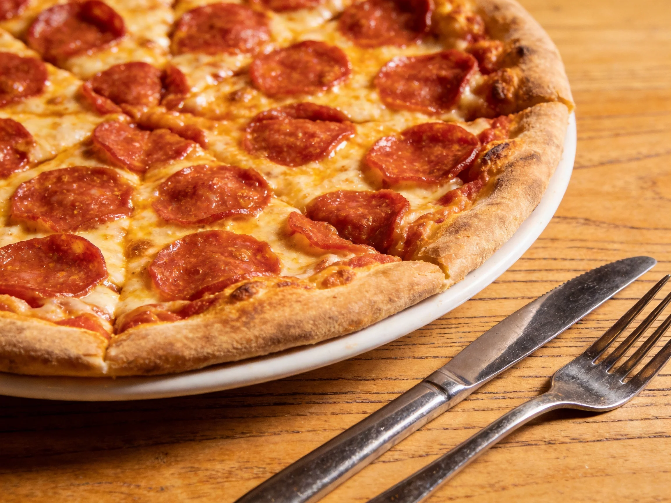 A large pepperoni pizza with golden crust on a white plate beside silverware