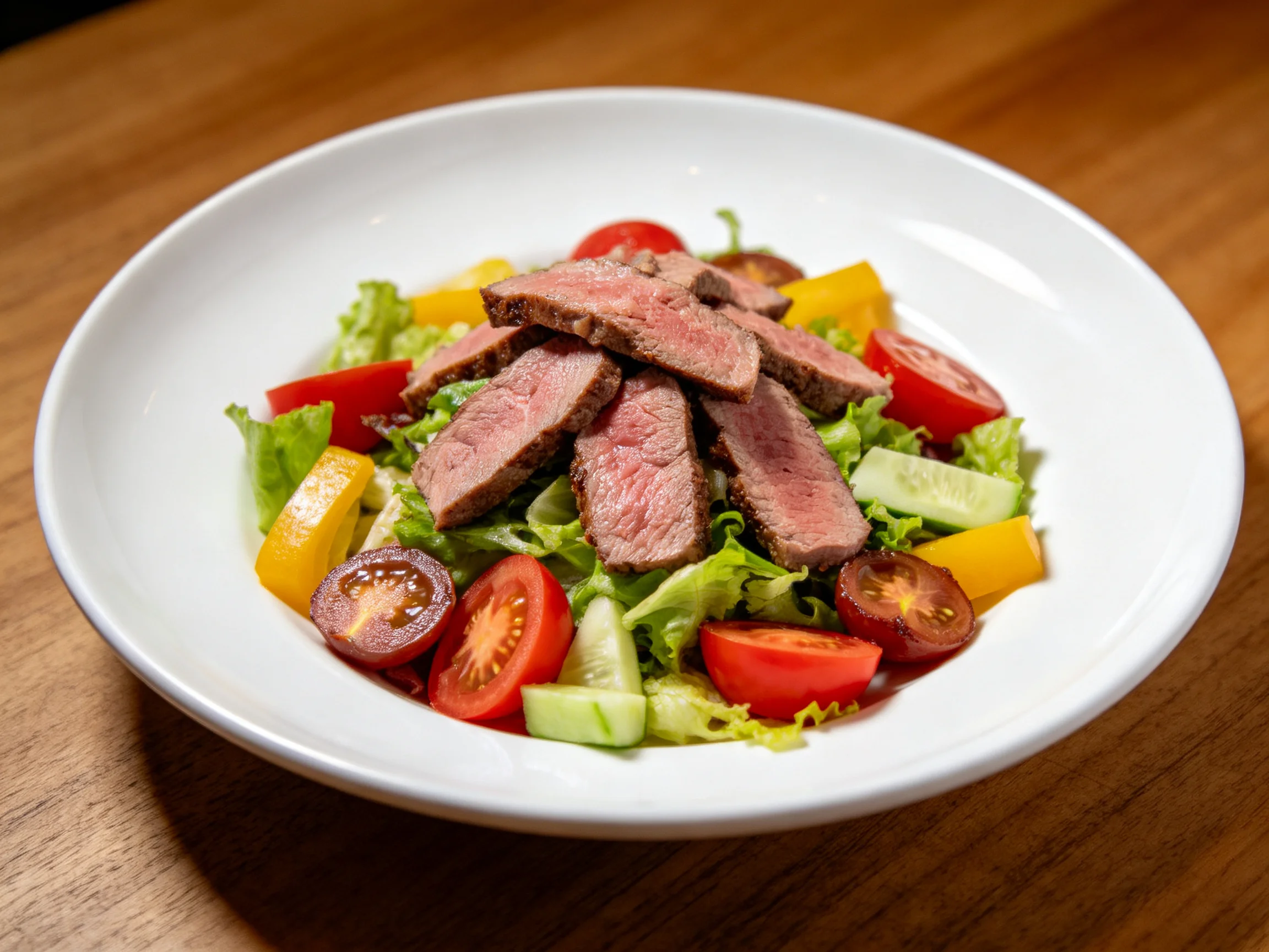 A white bowl holds a fresh salad with sliced steak, cherry tomatoes, and bell peppers