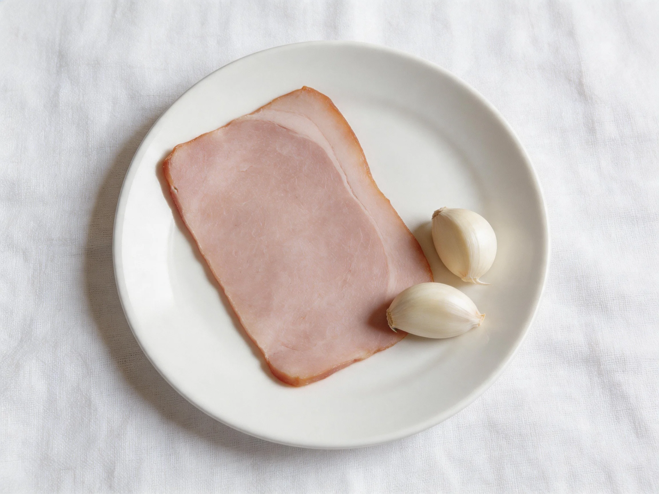 A white plate holds a slice of pink ham and two garlic cloves