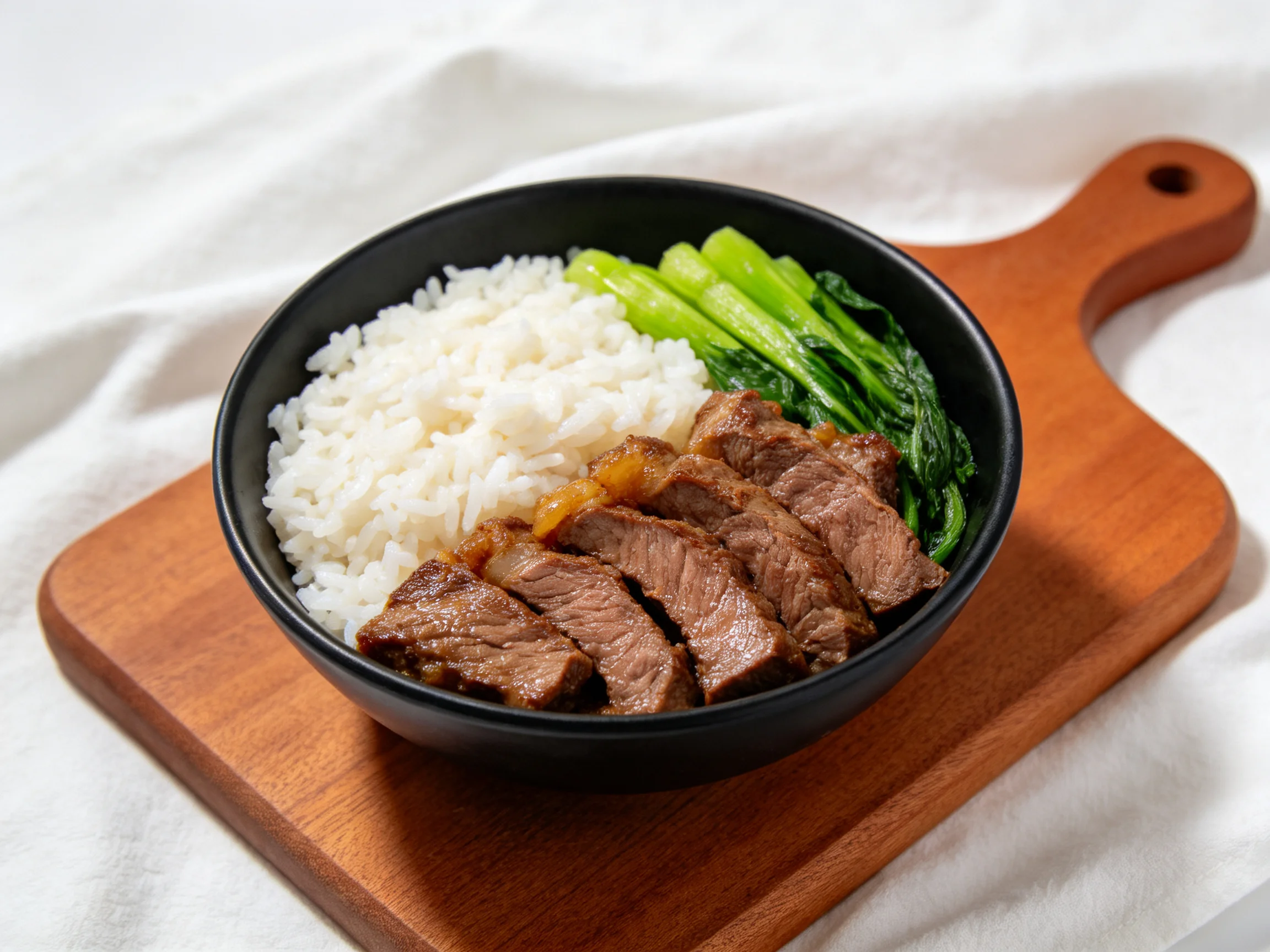 Savory beef slices, white rice, and greens in a black bowl on a wooden board