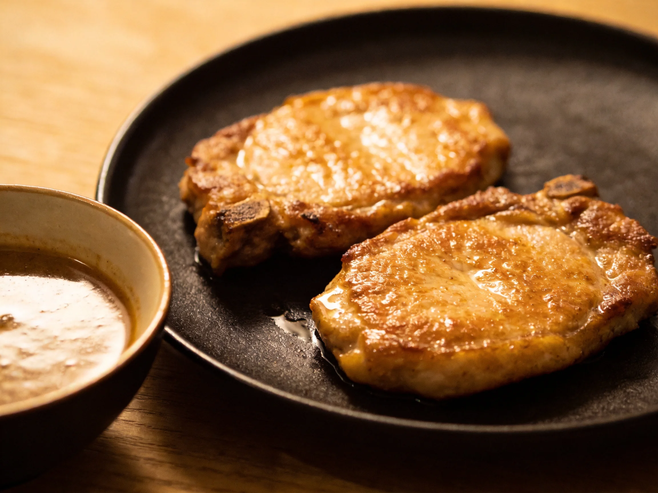 Two golden-brown pork cutlets served on a dark plate with a brown sauce bowl