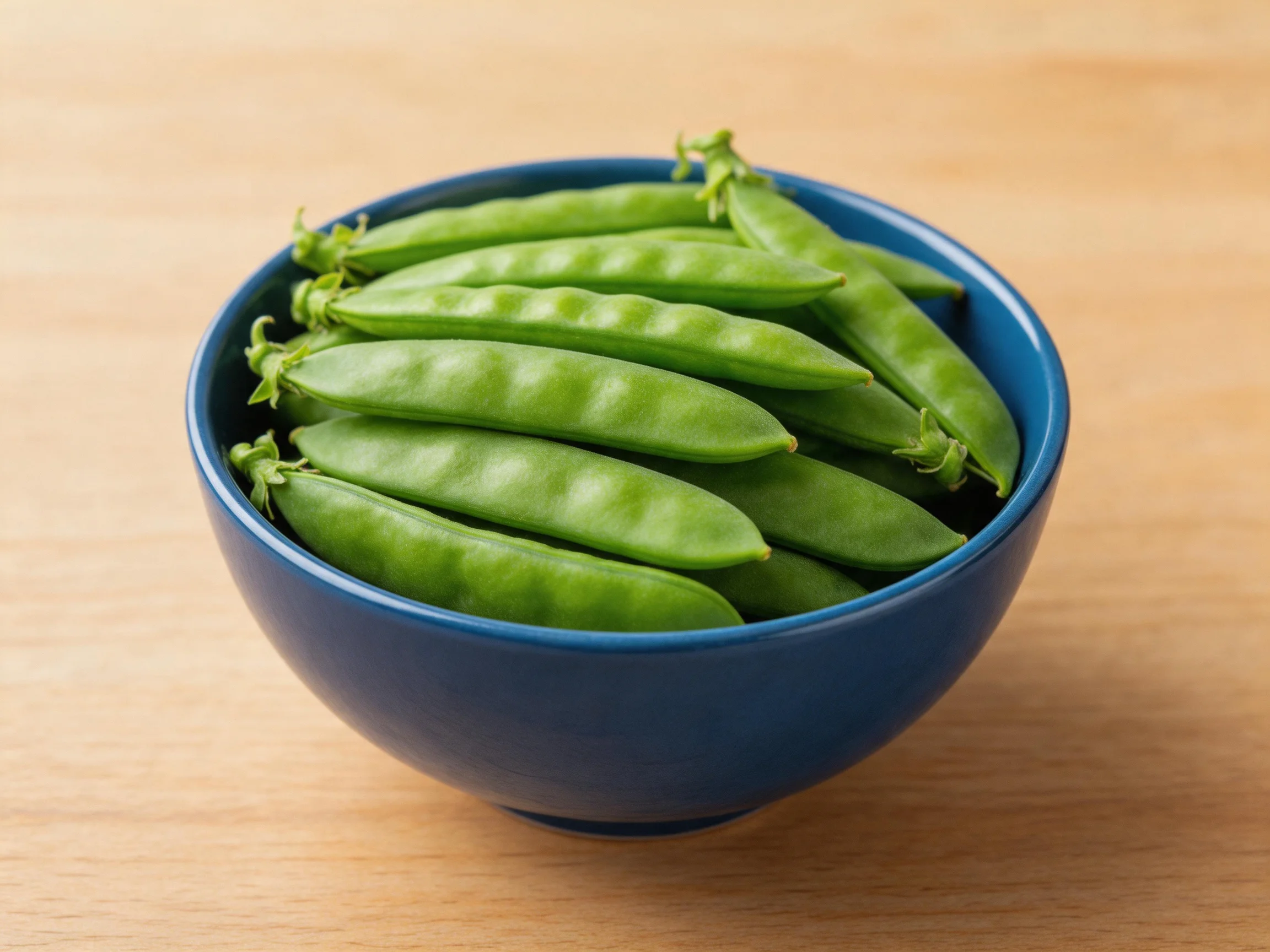 A blue ceramic bowl filled with fresh green sugar snap peas on a light wood surface